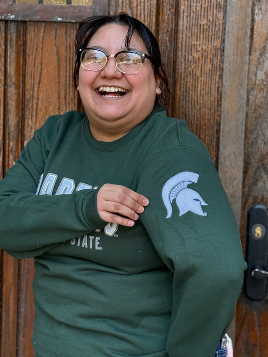 Close up of a woman wearing a green crewneck with "Spartans" written in white across the front and smaller white text underneath that reads "Michigan State." On the left sleeve is a white Spartan Helmet logo.