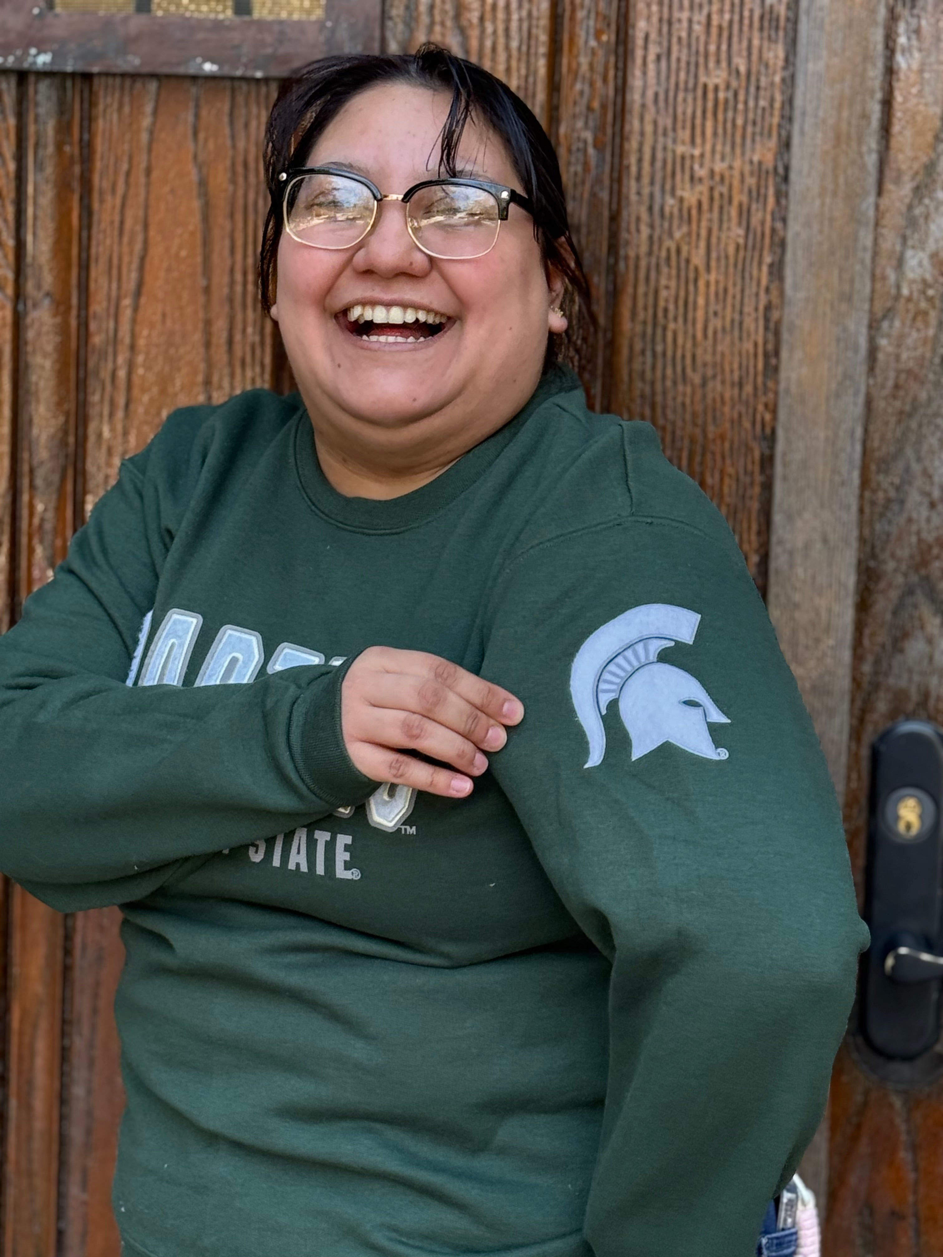Close up of a woman wearing a green crewneck with "Spartans" written in white across the front and smaller white text underneath that reads "Michigan State." On the left sleeve is a white Spartan helmet logo.