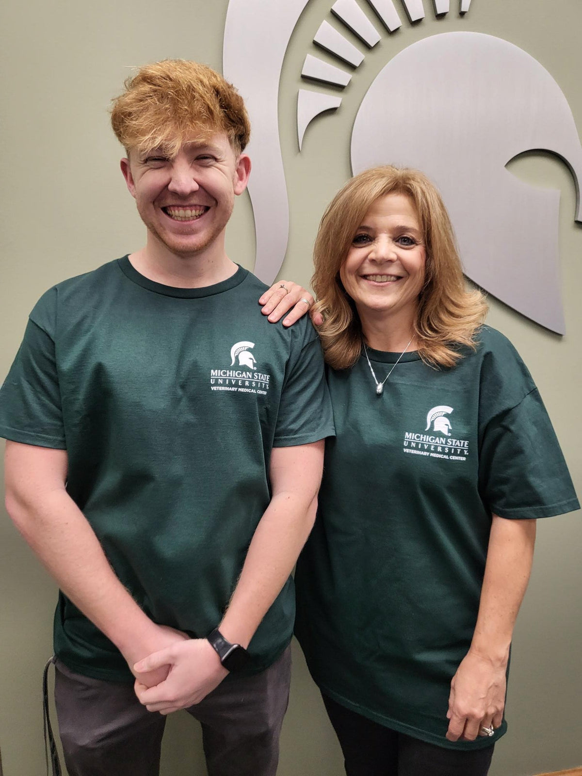 Two people with green T-shirts, featuring the Veterinary Medical Center logo and Spartan Helmet in white on the left upper chest.