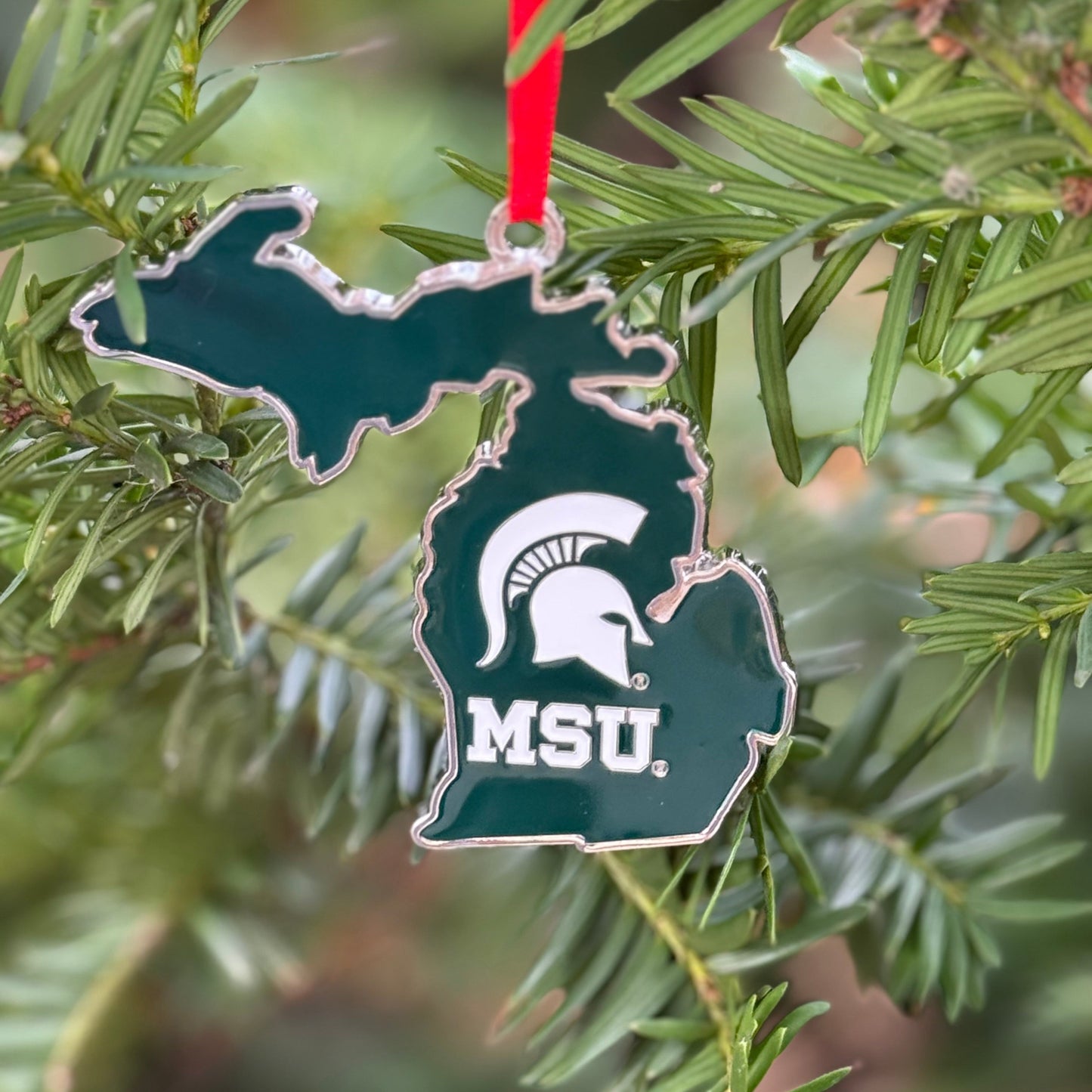 Green metal ornament shaped like the state of Michigan with a Spartan Helmet logo above the letters MSU in white. Shown hanging on a tree.