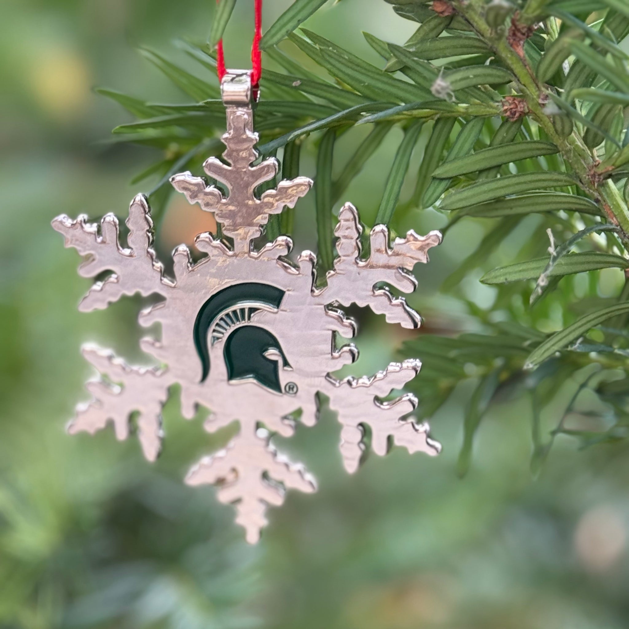 Silver snowflake-shaped ornament with a green Spartan helmet logo stamped in the centered of snowflake.