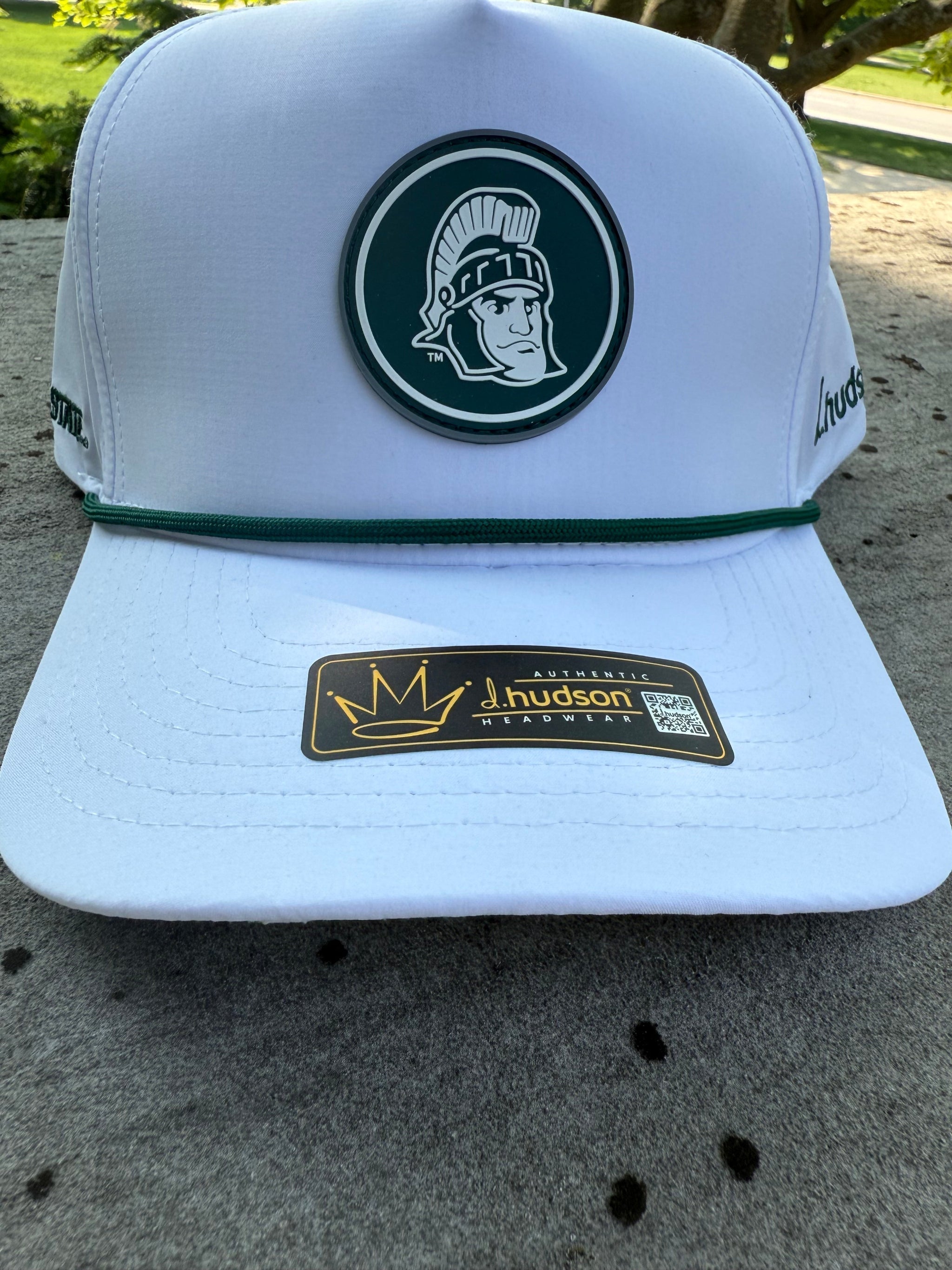 White hat with green cord on front, Green and white Sparty logo patch on the front, the words "Michigan State' embroidered in green on one side, and the brand name 'd.hudson' embroidered on the other side.