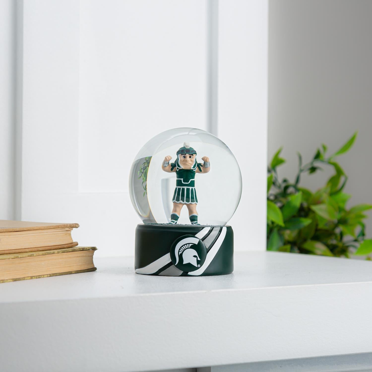 Sparty snow globe with a green base and Sparty figurine inside. The base has two white stripes, one gray stripe in the middle, and a Spartan helmet logo in the middle of the the stripes.