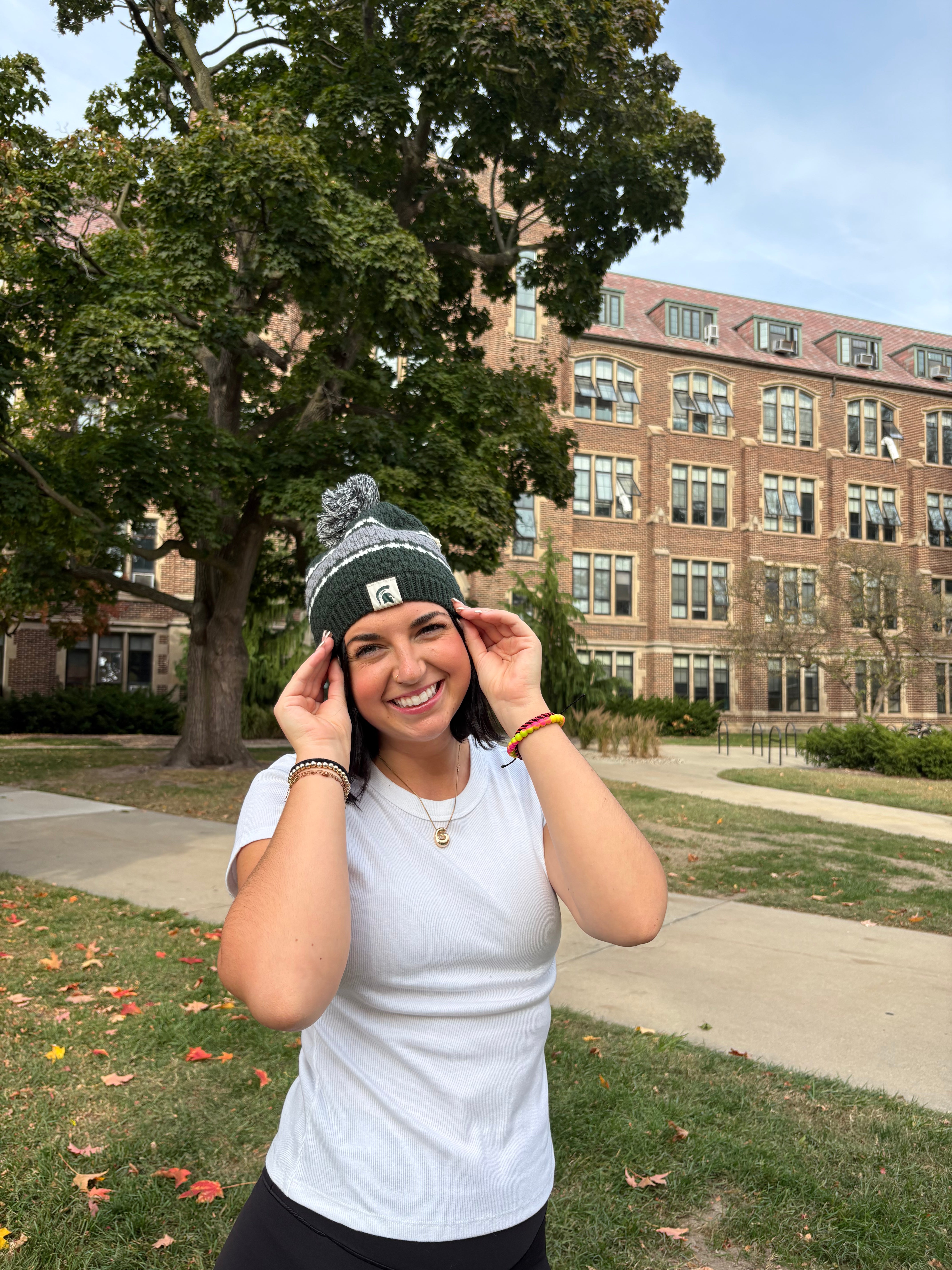 Woman standing outdoors, wearing a Michigan State themed green beanie with a thick gray stripe and two thin white stripes around the middle of the hat. The beanie has a green and white pom an the top and a Spartan helmet patch sewn on the cuff.