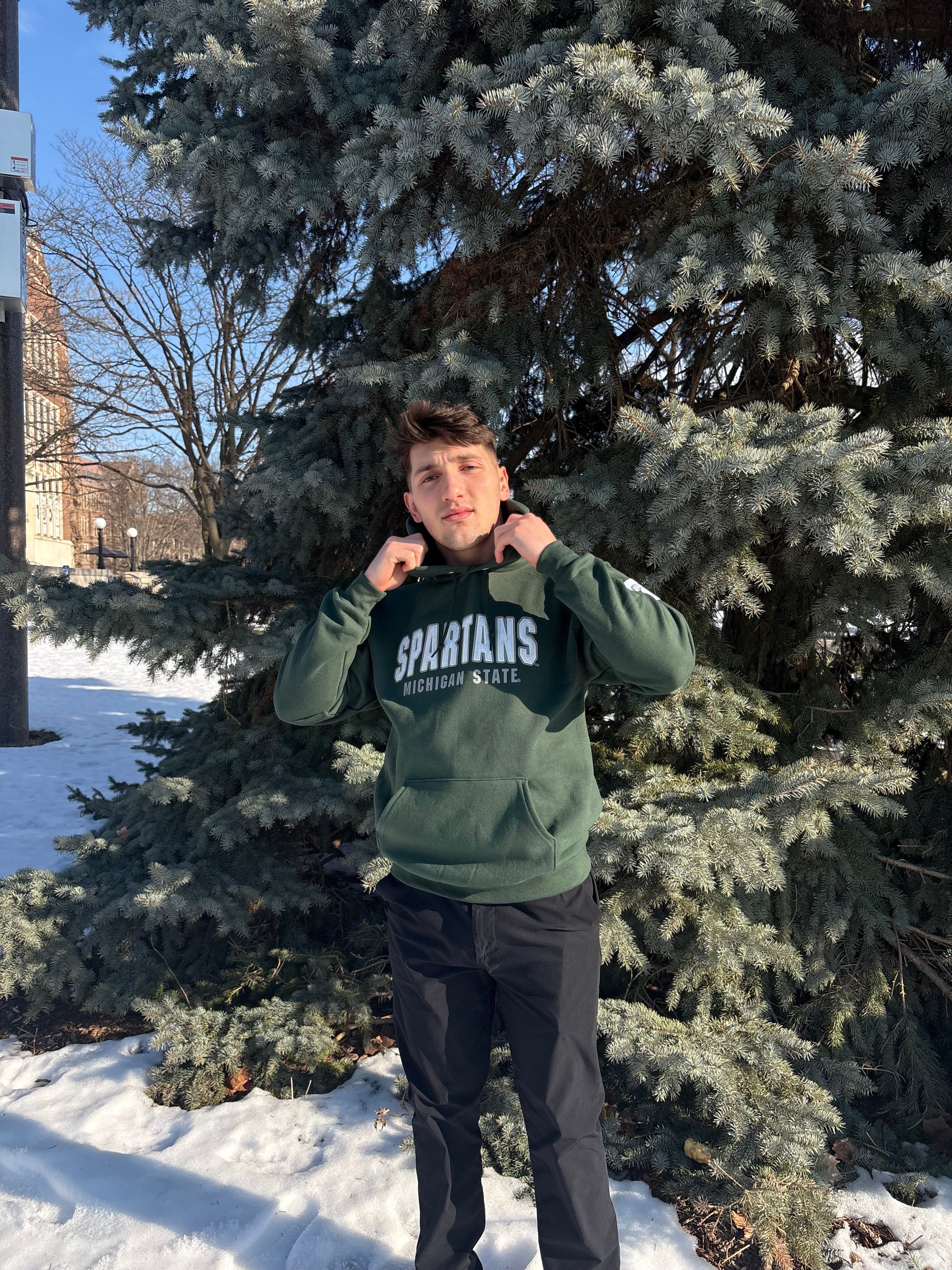 Man standing outdoors, wearing a green hoodie with "Spartans" written across the front in white and smaller white text underneath that reads "Michigan State."