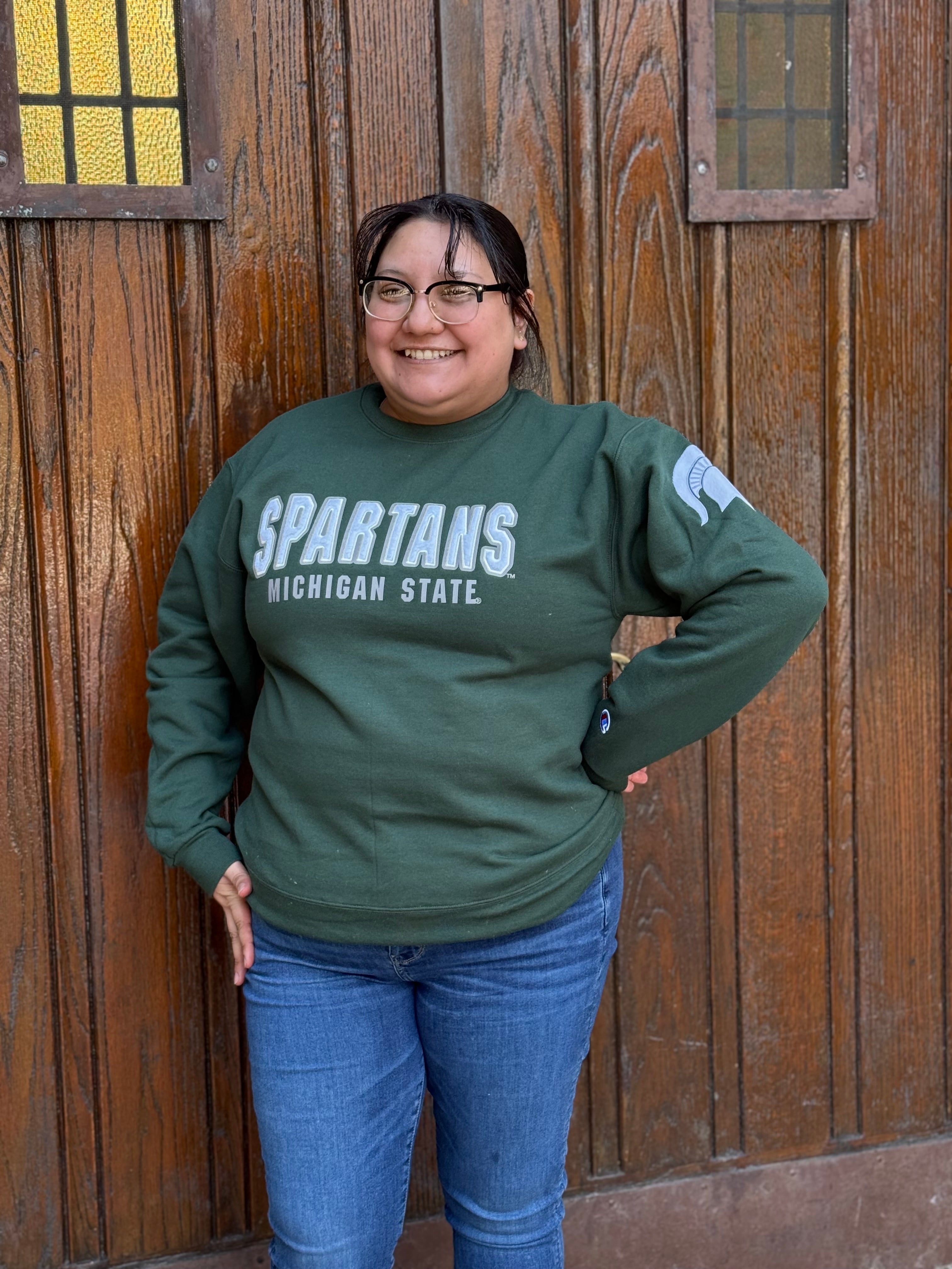 Woman wearing a green crewneck with "Spartans" written in white across the front and smaller white text underneath that reads "Michigan State." On the left sleeve is a white Spartan helmet logo.