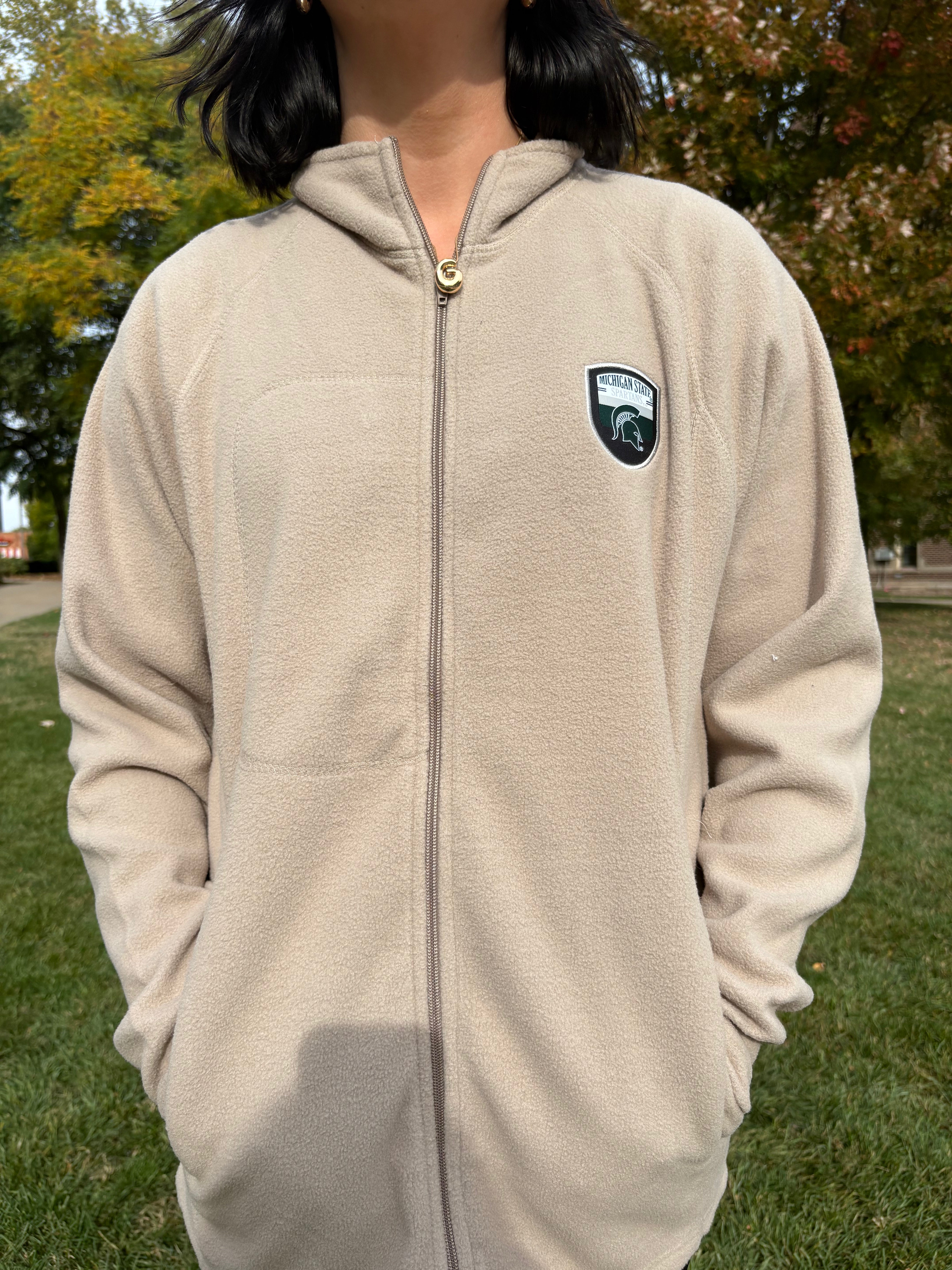 Person wearing a beige fleece jacket with a logo on the chest outdoors.