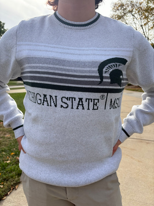 Close up of a person wearing a light gray sweater with white, gray, and black stripes on the center chest section and a 'Michigan State \ MSU' wordmark in dark green below. There is a big dark green Spartan Helmet logo on the left upper chest and a dark green stripe on the collar, sleeve, and bottom hems.
