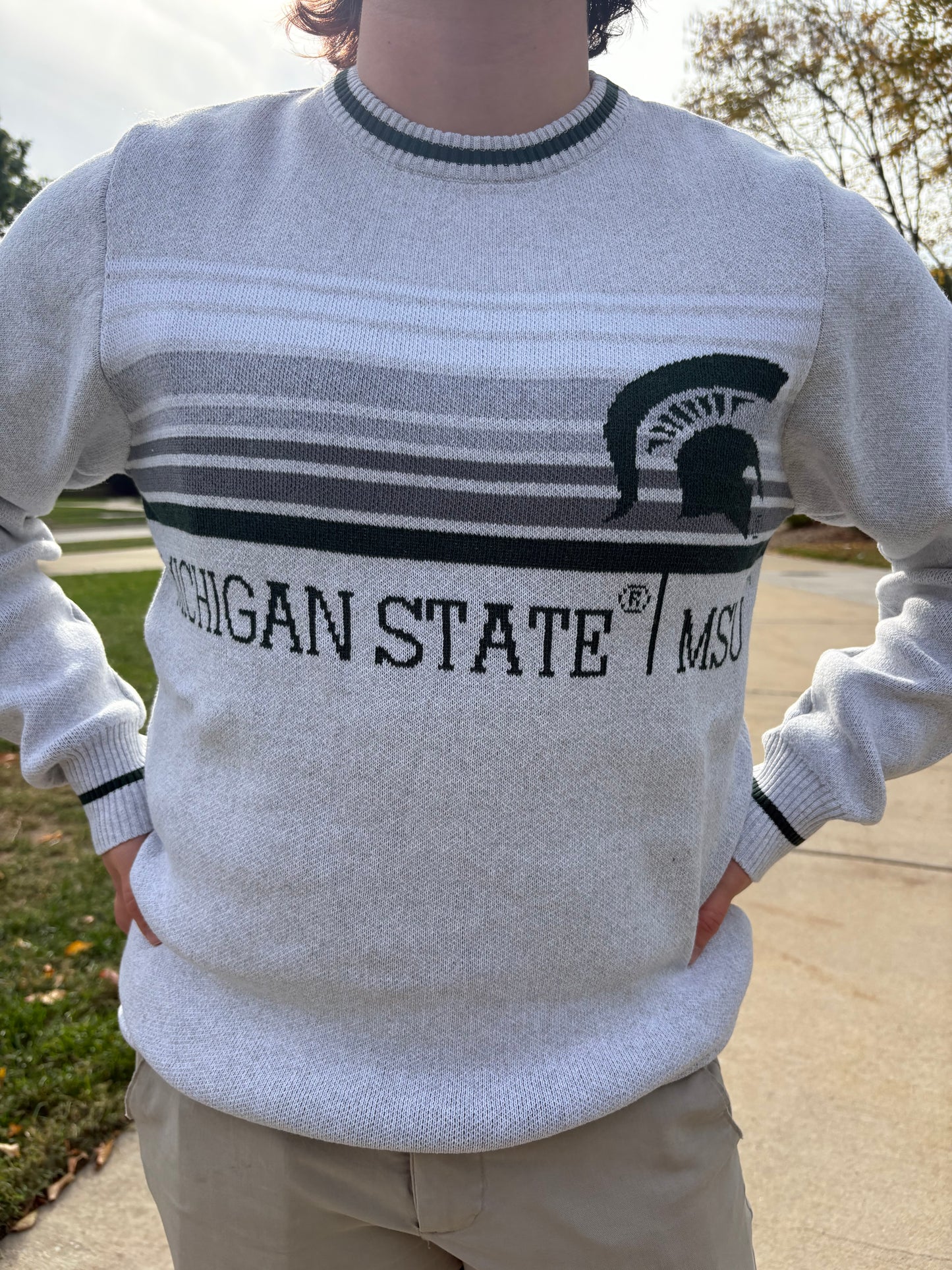 Close up of a person wearing a light gray sweater with white, gray, and black stripes on the center chest section and a 'Michigan State \ MSU' wordmark in dark green below. There is a big dark green Spartan Helmet logo on the left upper chest and a dark green stripe on the collar, sleeve, and bottom hems.