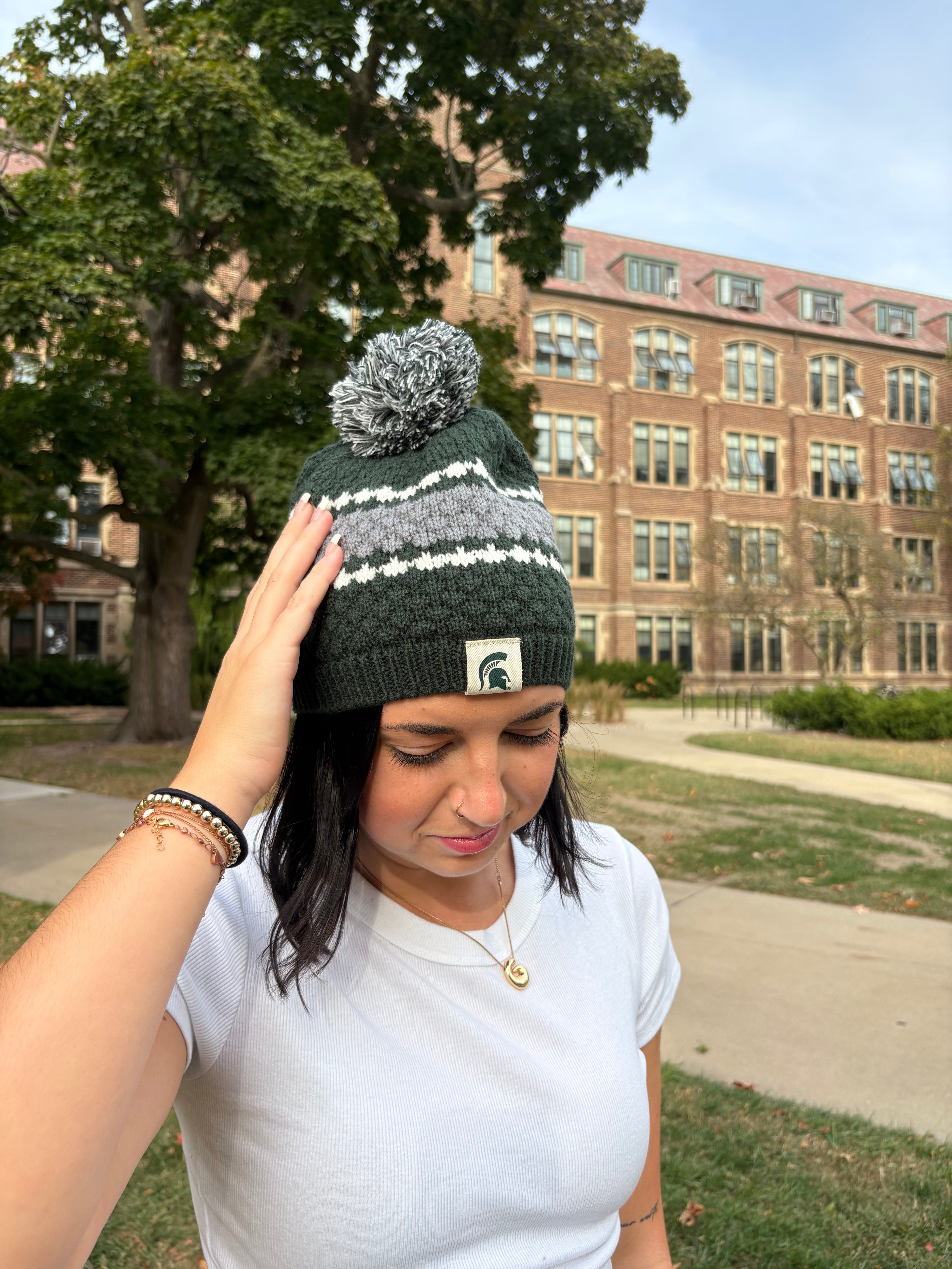 Closer up image of a woman standing outdoors, wearing a Michigan State themed green beanie with a thick gray stripe and two thin white stripes around the middle of the hat. The beanie has a green and white pom an the top and a Spartan helmet patch sewn on the cuff.