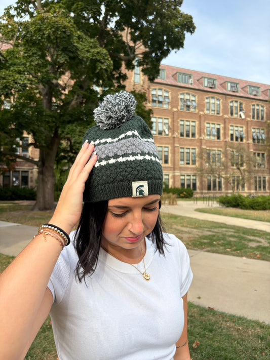 Closer up image of a woman standing outdoors, wearing a Michigan State themed green beanie with a thick gray stripe and two thin white stripes around the middle of the hat. The beanie has a green and white pom an the top and a Spartan Helmet patch sewn on the cuff.