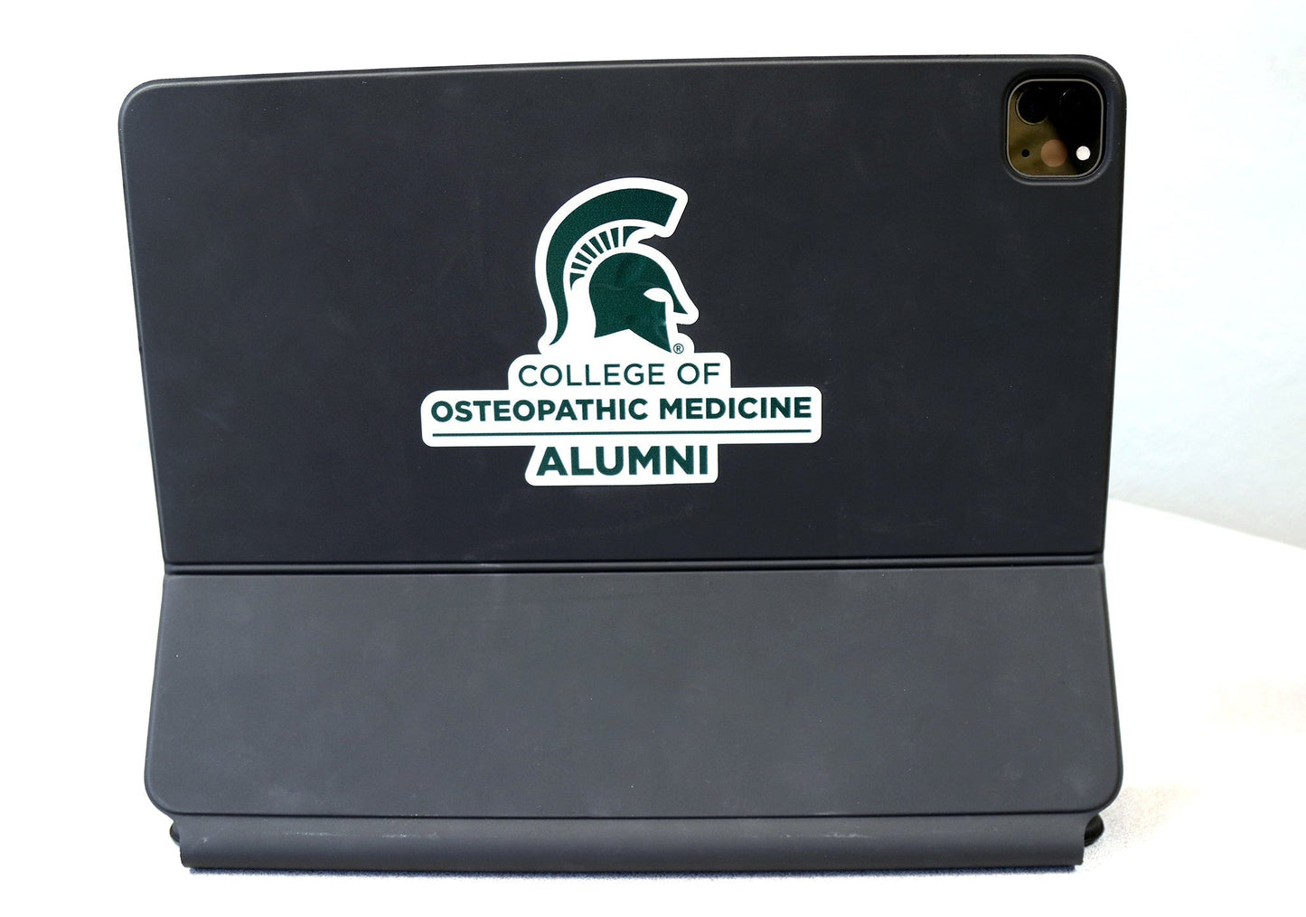 Back of black laptop with a white decal with the Spartan Helmet and College of Osteopathic Medicine Alumni in forest green.