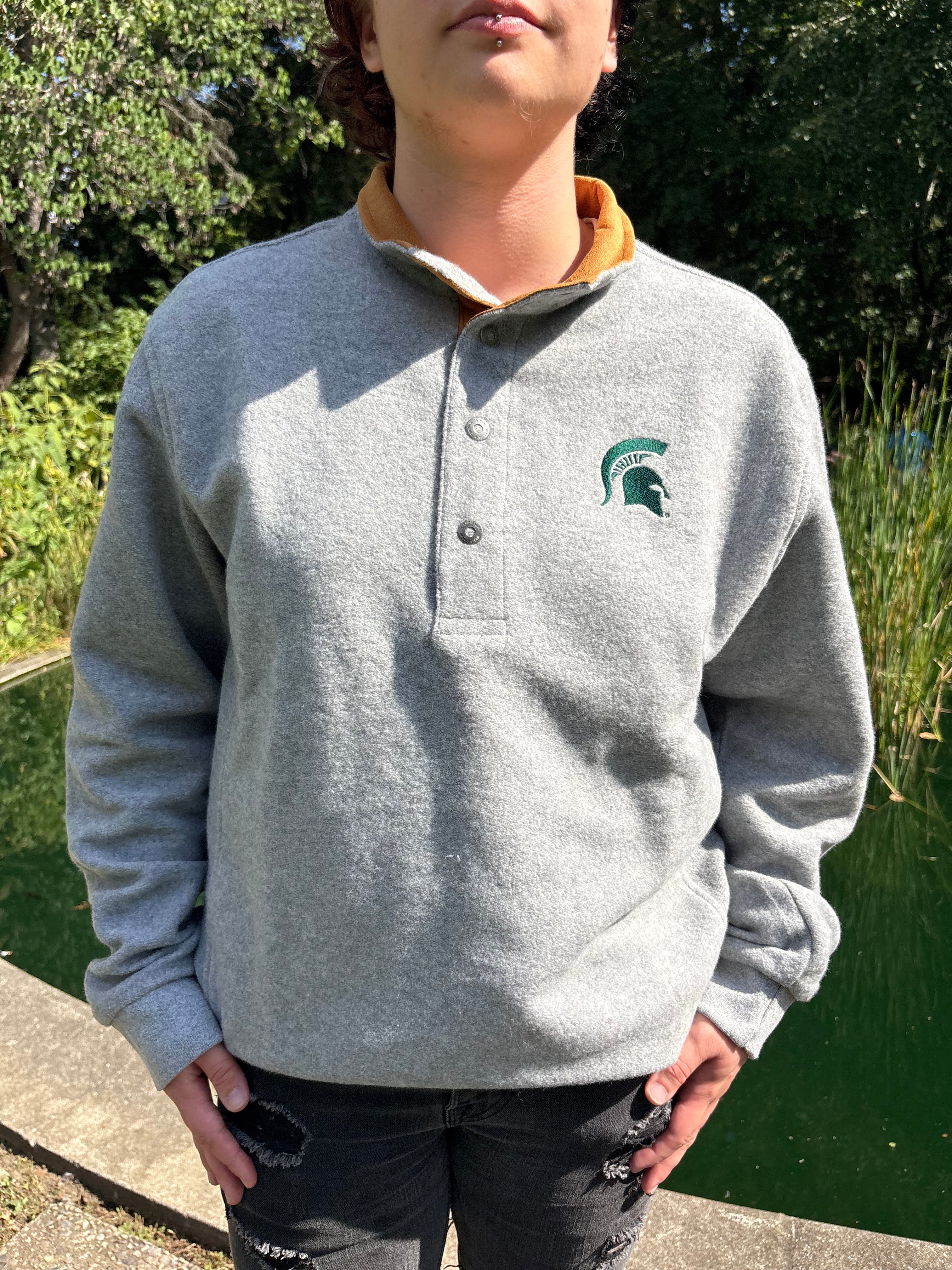 Person standing by a body of water, wearing a button-up quarter-zip gray jacket with orange contrasting on the collar and a green embroidered Spartan helmet logo on the left upper chest.