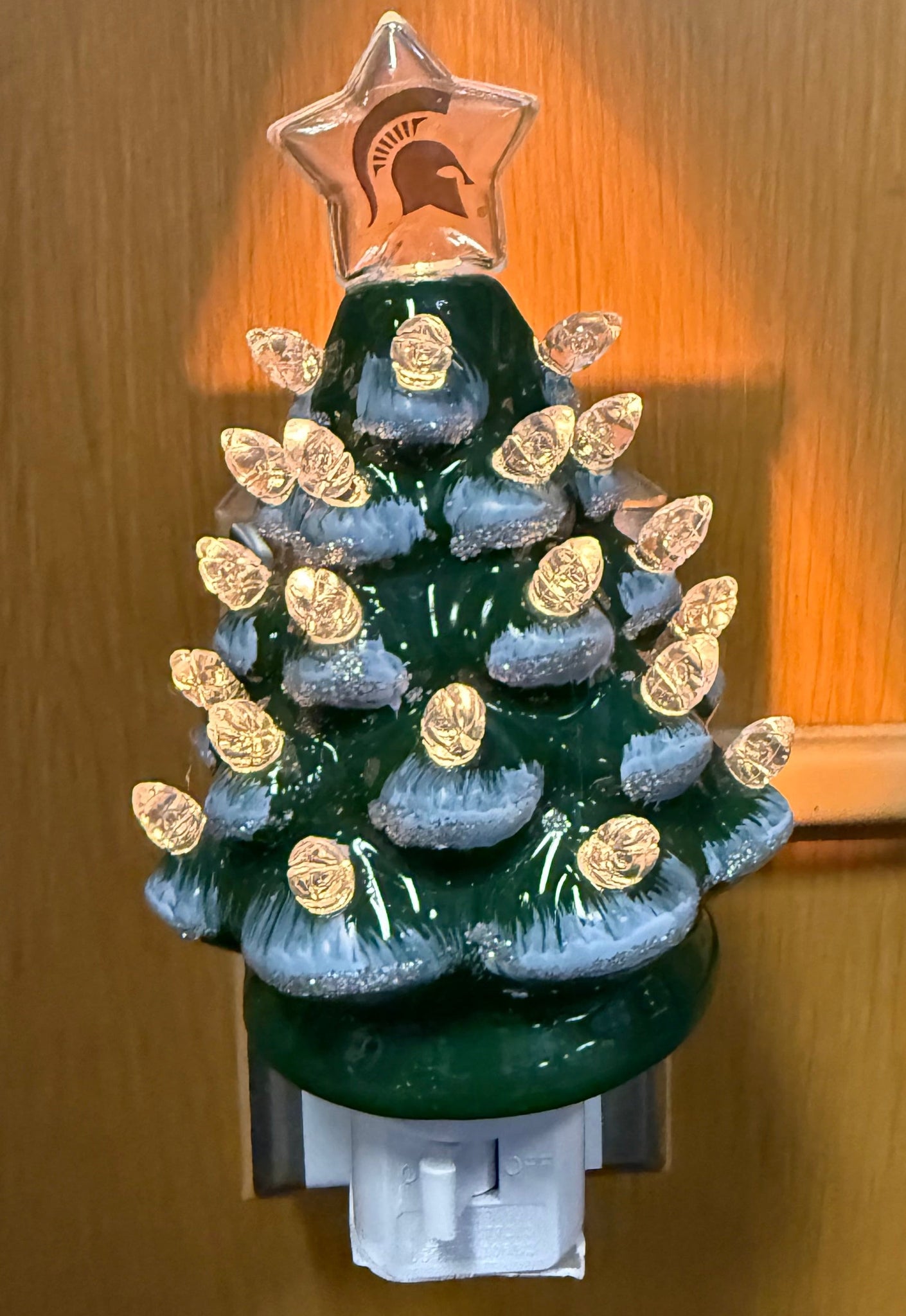 Ceramic tree night light in a evergreen tree shape with a glossy green finish, accented by painted snow tipped branches and clear bulb-style ornaments. A clear start with a green Spartan helmet logo on it is on top of the tree night light. There is a white on and off switch at the bottom of the night light. Shown plugged in and lit up.