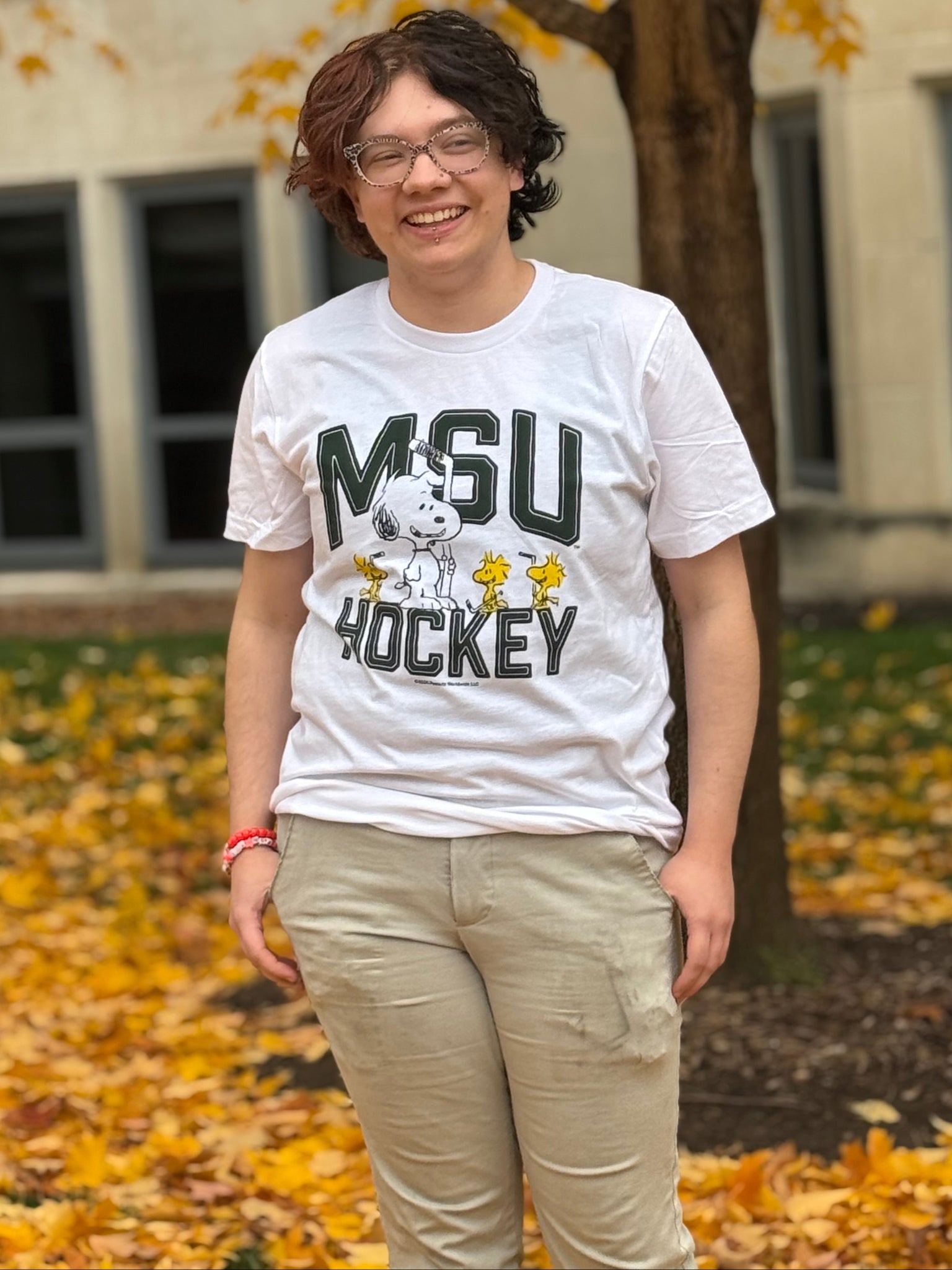 Person standing outdoors in front of a tree with yellow autumn leaves, wearing a white T-shirt with ‘MSU Hockey’ text, a Snoopy graphic, and light-colored pants.