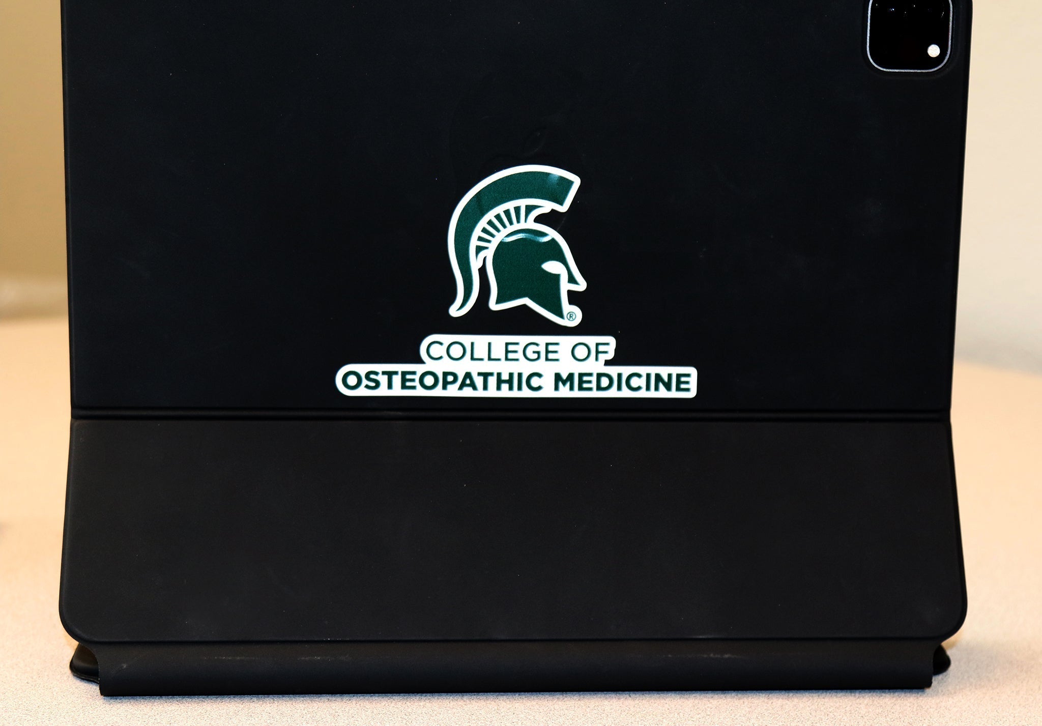 White vinyl transfer decal with a forest green College of Osteopathic Medicine logo and Spartan helmet logo on it. Shown on top of a black iPad case.
