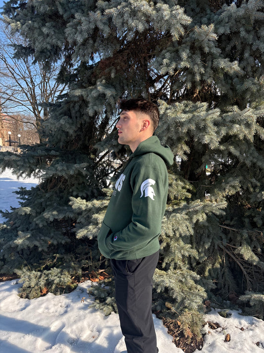 Man standing outdoors, showing the left side of a green hoodie. The left sleeve has a white Spartan Helmet logo.