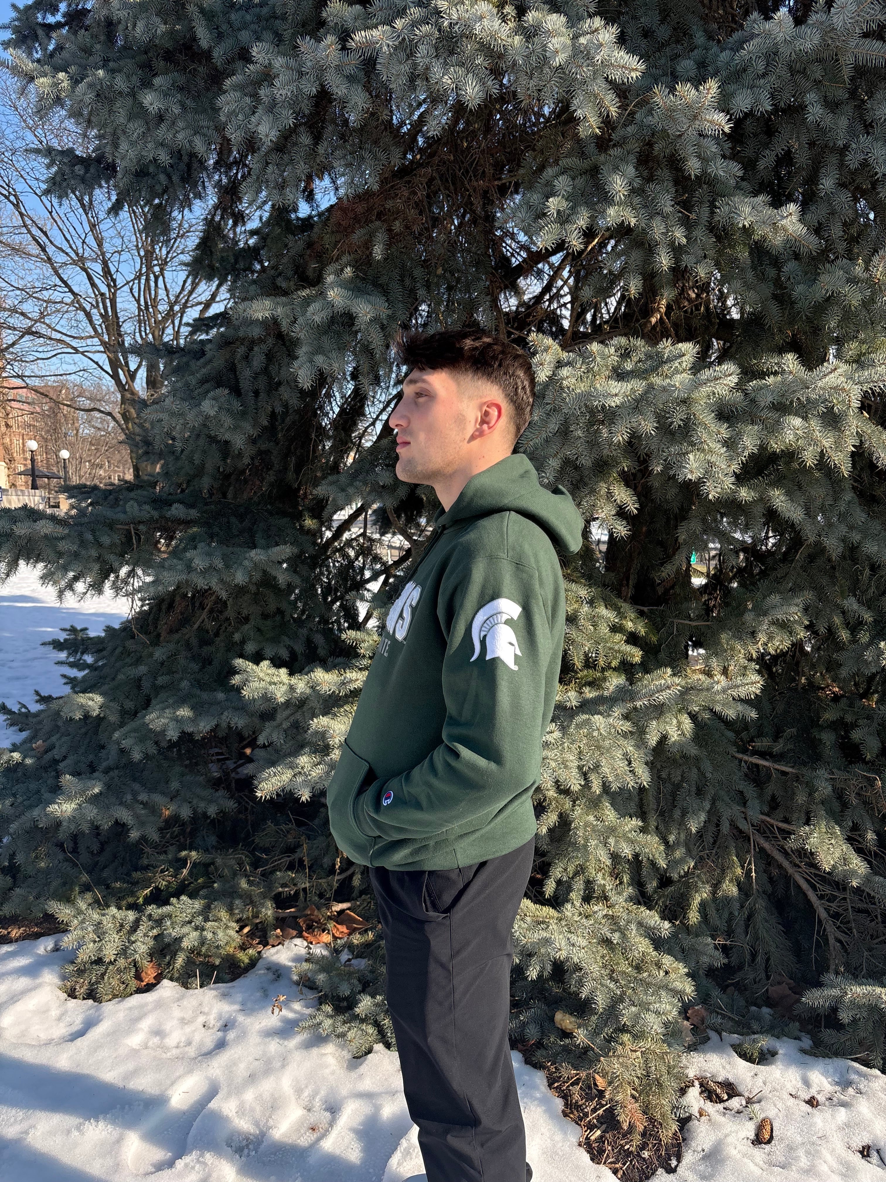 Man standing outdoors, showing the left side of a green hoodie. The left sleeve has a white Spartan helmet logo.
