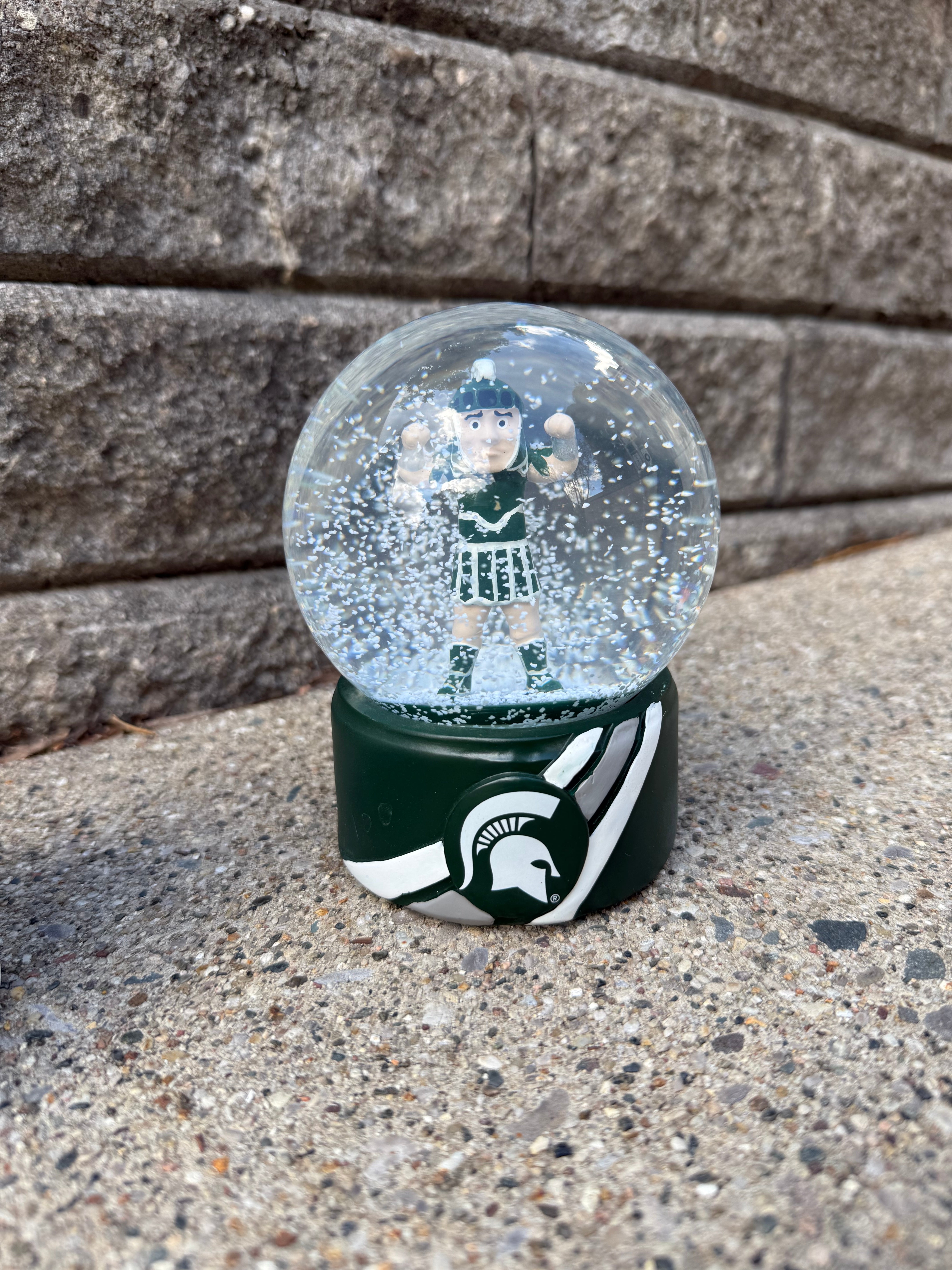 Sparty snow globe with a green base and Sparty figurine inside. The base has two white stripes, one gray stripe in the middle, and a Spartan helmet logo in the middle of the the stripes.