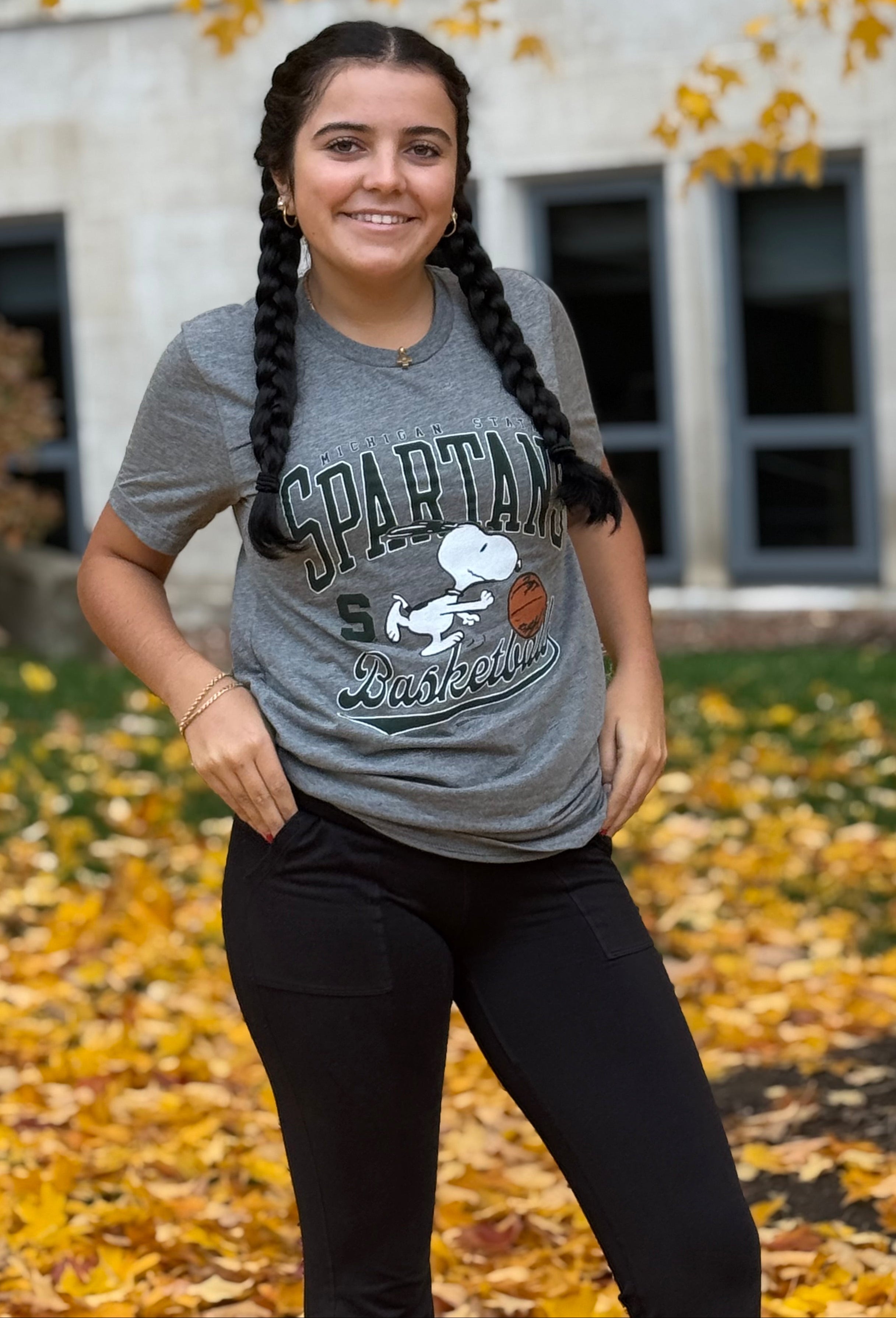 Person standing outdoors in front of a building and tree with yellow autumn leaves, wearing a gray T-shirt with the text ‘Spartans Basketball’ and a Snoopy graphic that is screen printed, paired with black pants. Fallen leaves cover the ground.