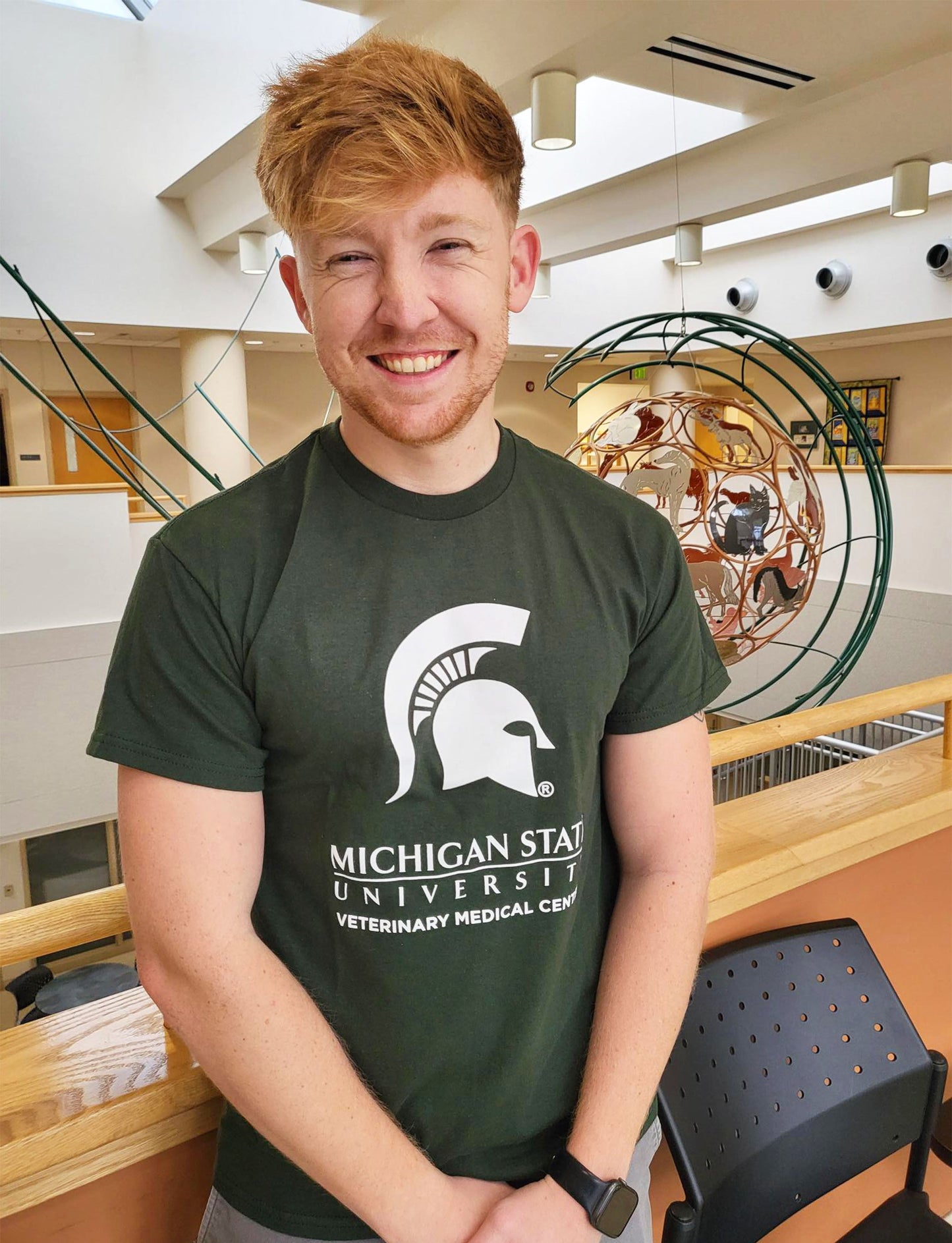 A man in blue jeans wearing a green short sleeve T-shirt. Across the center chest is a large white Spartan Helmet centered above the Michigan State wordmark logo. Beneath that is large block text reading “Veterinary Medical Center.”