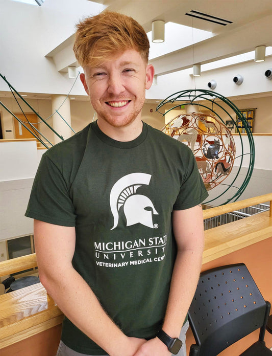 A man in blue jeans wearing a green short sleeve T-shirt. Across the center chest is a large white Spartan helmet centered above the Michigan State wordmark logo. Beneath that is large block text reading “Veterinary Medical Center.”