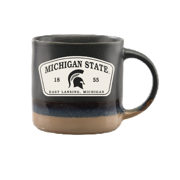 Michigan State Spartans ceramic mug featuring a black and tan reactive glaze finish with a large white badge-style graphic displaying “Michigan State,” the Spartan helmet logo, “1855,” and “East Lansing, Michigan.” The mug has a curved handle and a two-tone handcrafted appearance with a dark upper section and natural stoneware-colored base.