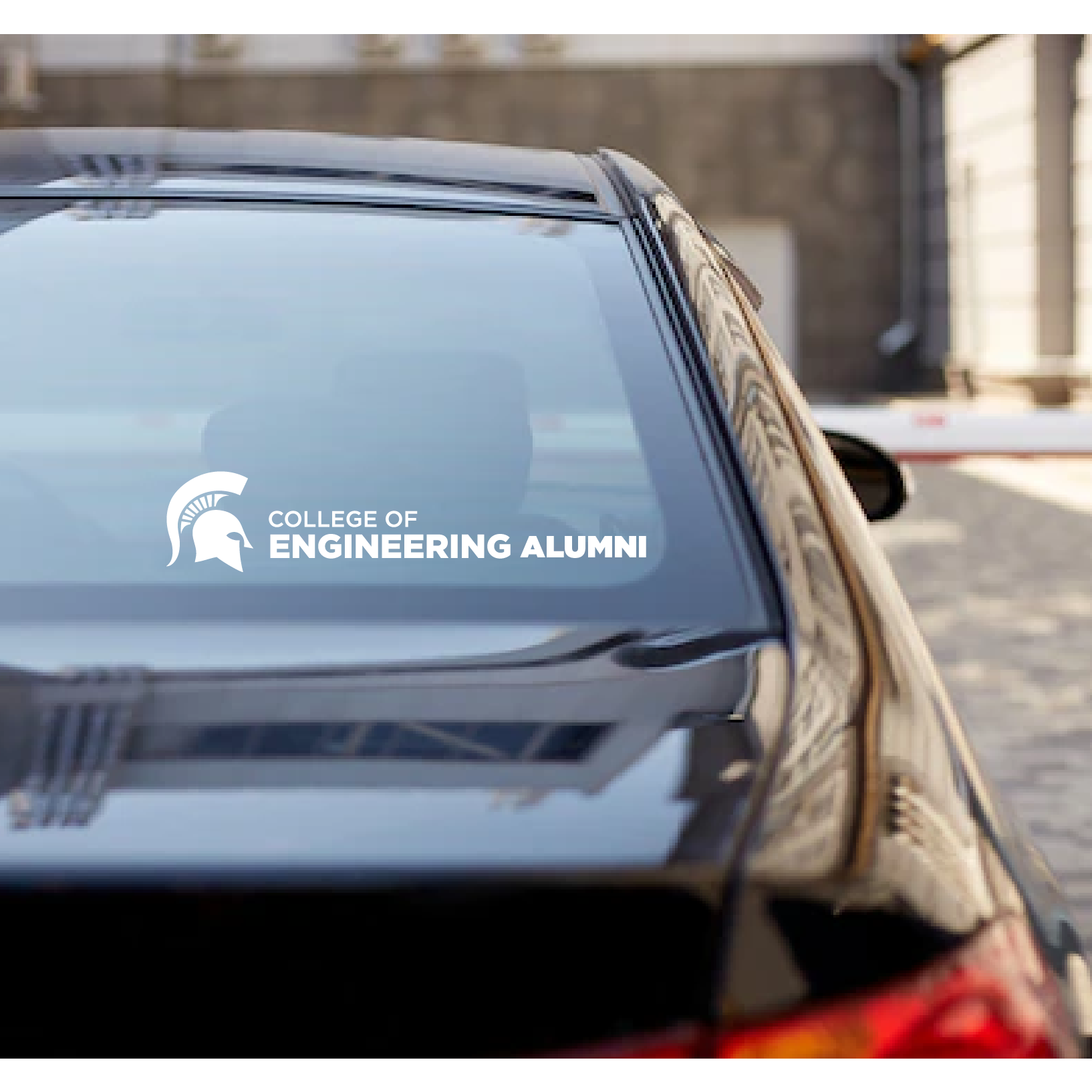 Decal reading College of Engineering Alumni in all caps next to a white Spartan helmet on the back rear windshield of a dark sedan.
