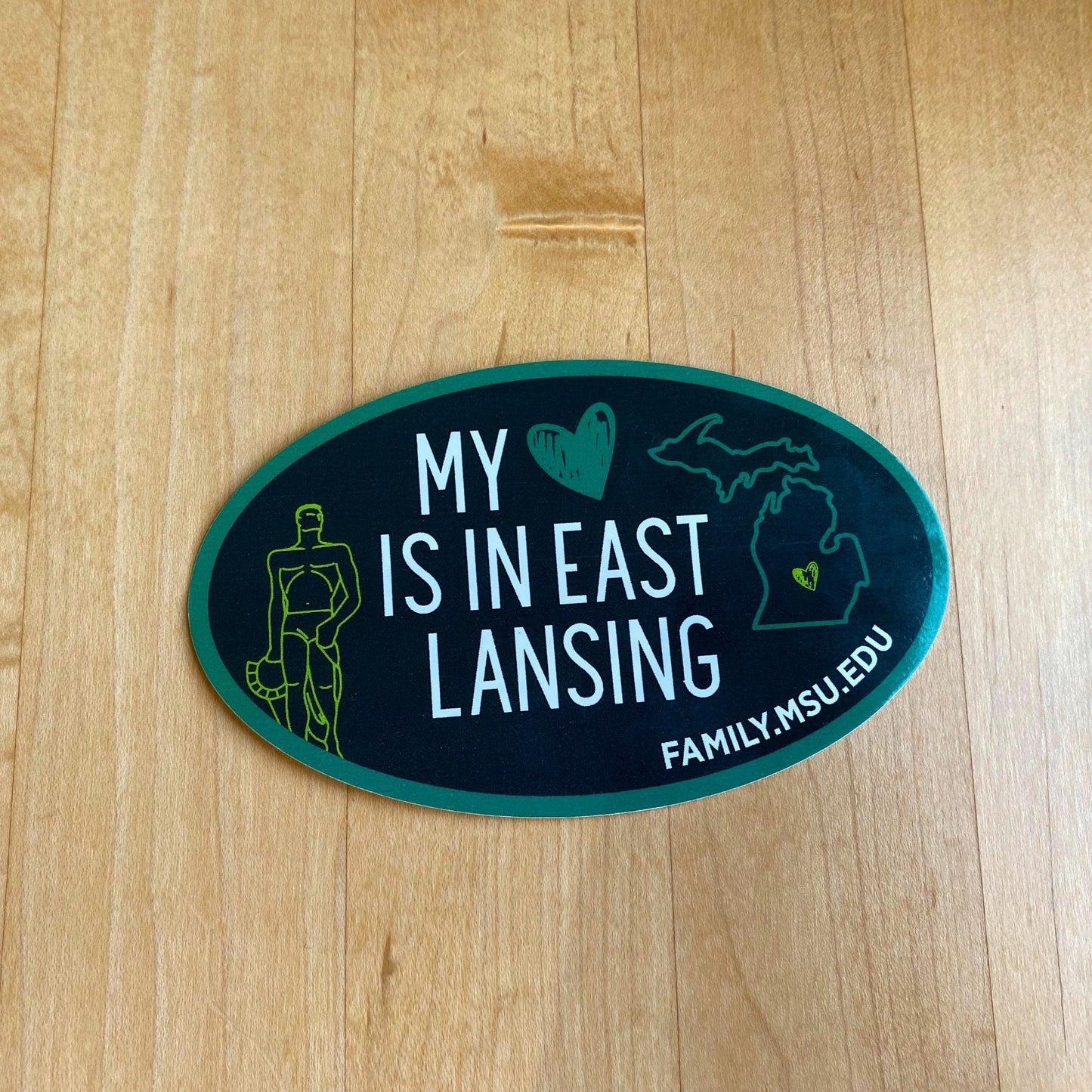 A green oval vinyl sticker with Spartan Statue and state of Michigan outlined in green with a green heart over East Lansing. In the center of the sticker it says, "My heart is in East Lansing" in white with a green heart next to it.