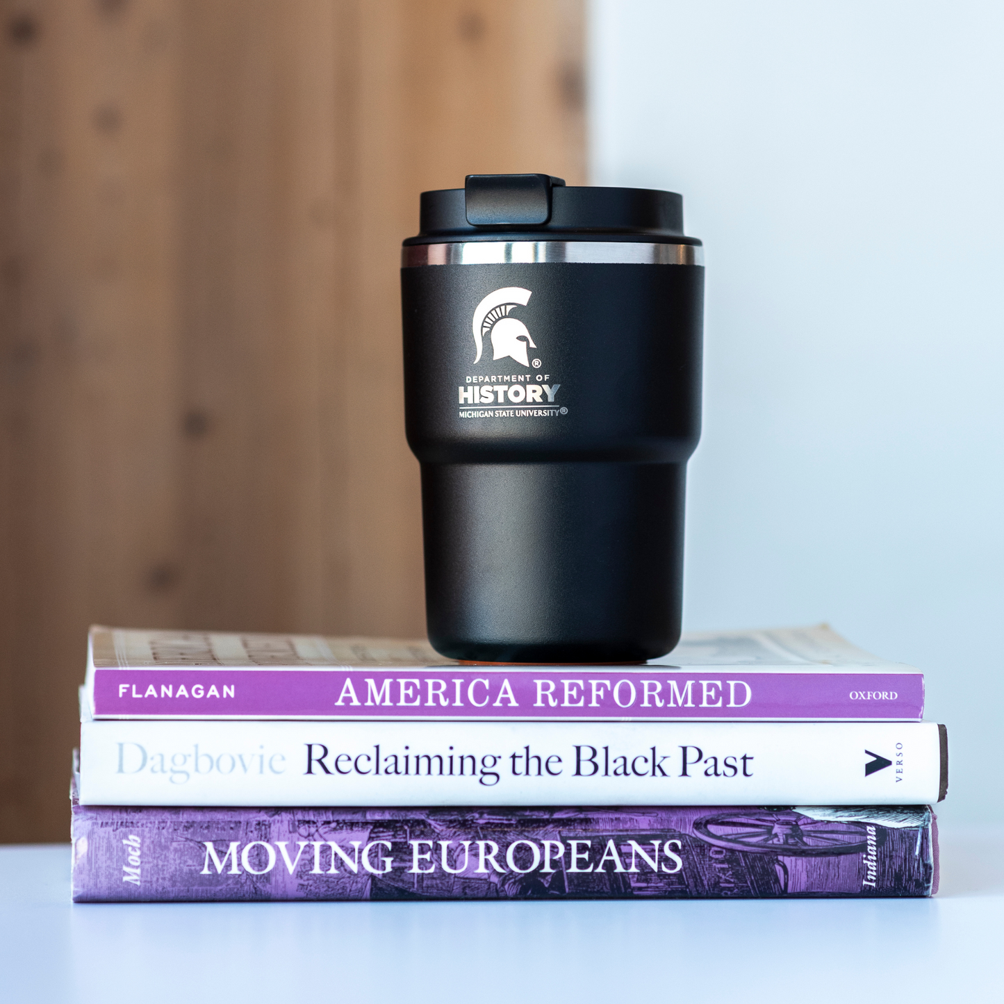 A black 12 ounce mug with a laser-etched white Department of History logo and Spartan Helmet sitting on top of 3 books.