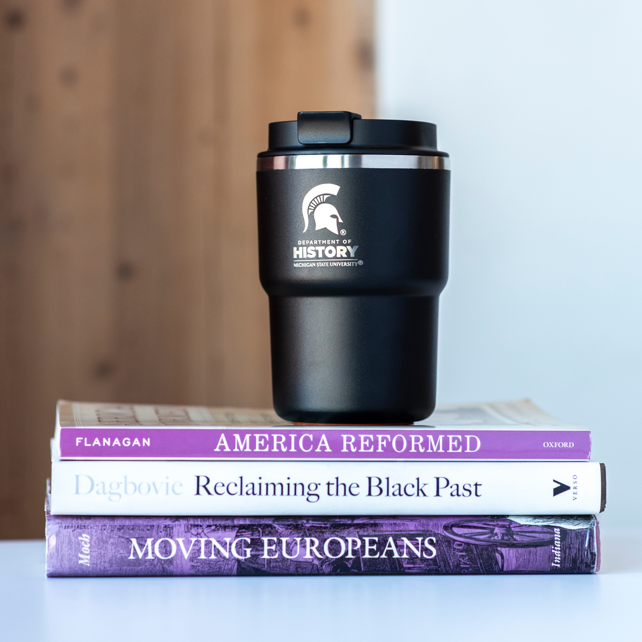 A black 12 ounce mug with a laser-etched white Department of History logo and Spartan helmet sitting on top of 3 books.