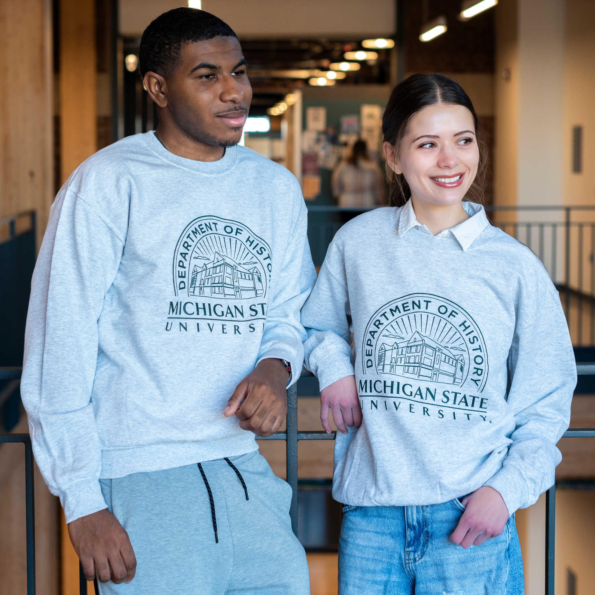 Two individuals wearing gray crewneck sweatshirts that showcases the Old Horticulture building surrounded by the text ‘Department of History’ and ‘Michigan State University’.