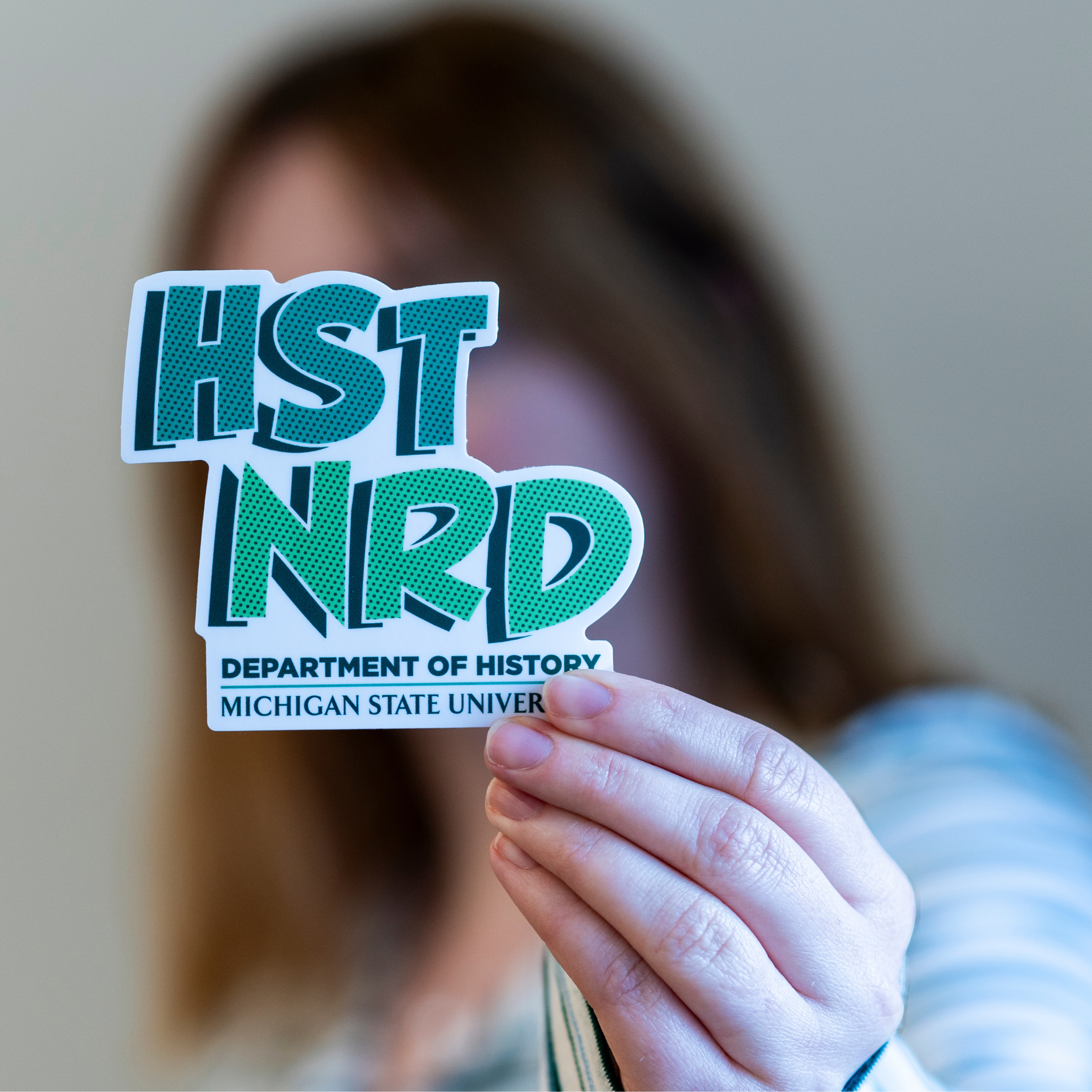 Person holding green and white vinyl sticker that features the text ‘HST NRD’ above of ‘Department of History’ and ‘Michigan State University’.