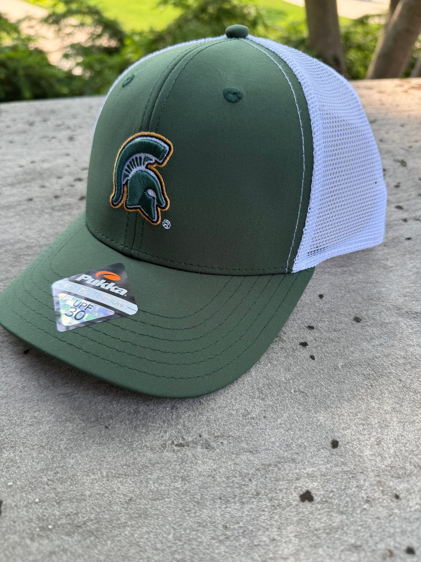 Green cap leaning against concrete outside featuring a Spartan Helmet logo in the middle and a white mesh back.