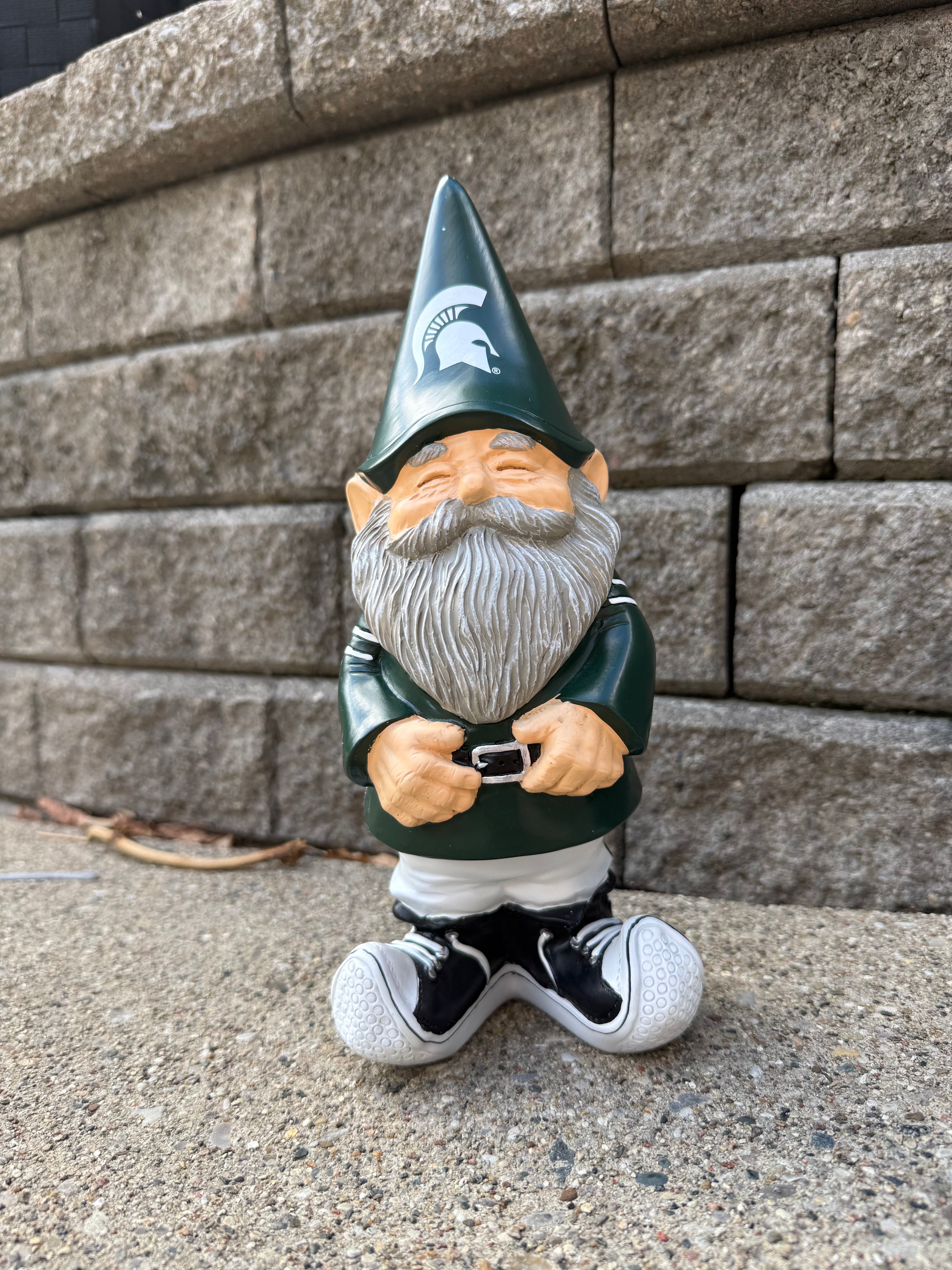 Garden gnome with a green hat and gray beard, standing on a concrete surface against a stone wall. The gnome's hat has a white Spartan helmet logo on it.