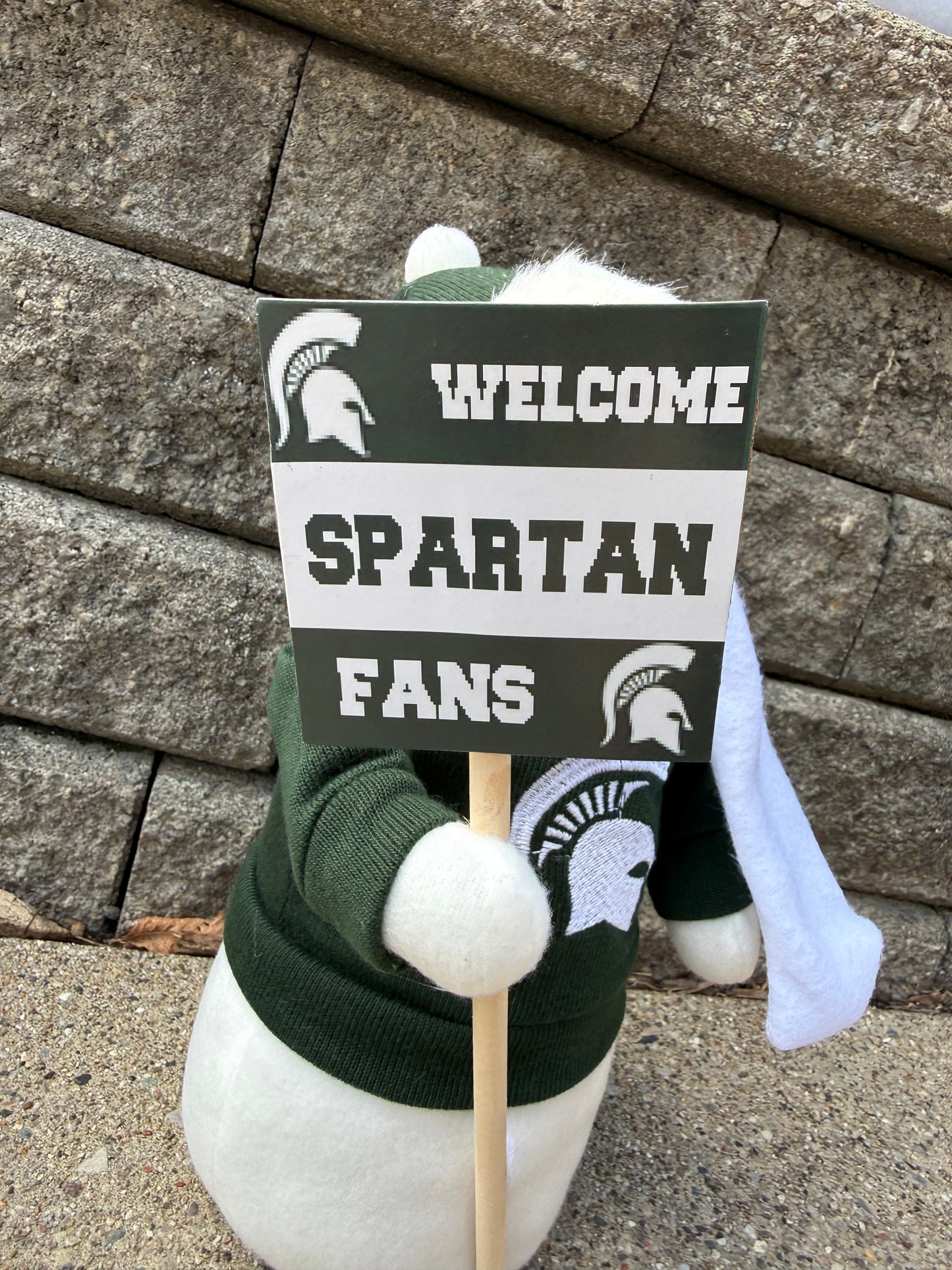 Close up of the snowman figurine wearing a winter hat, green sweater with a Spartan Helmet logo on it, and a white scarf. The snowman is holding a sign that says "Welcome Spartan Fans."