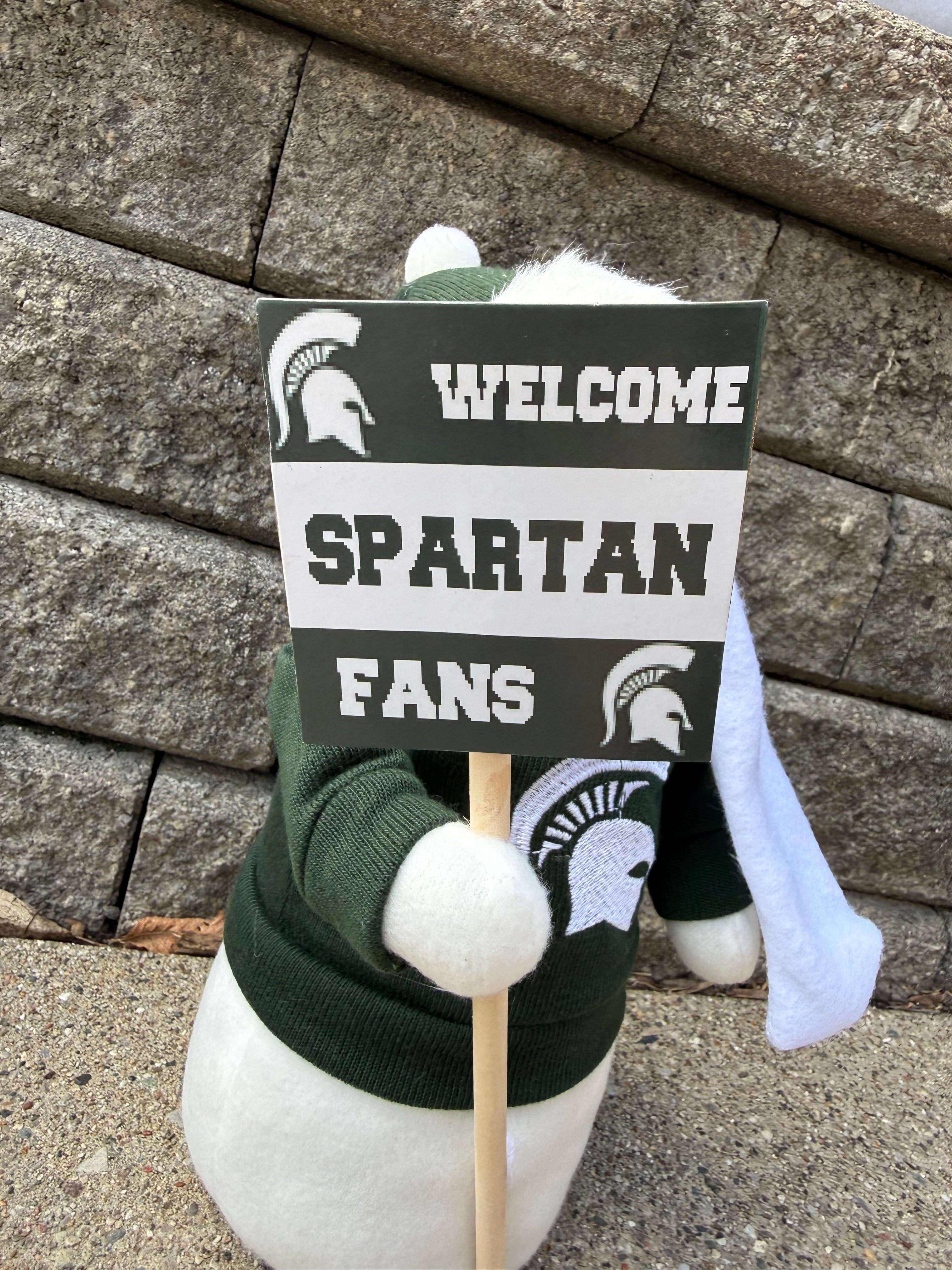 Close up of the snowman figurine wearing a winter hat, green sweater with a Spartan helmet logo on it, and a white scarf. The snowman is holding a sign that says "Welcome Spartan Fans".