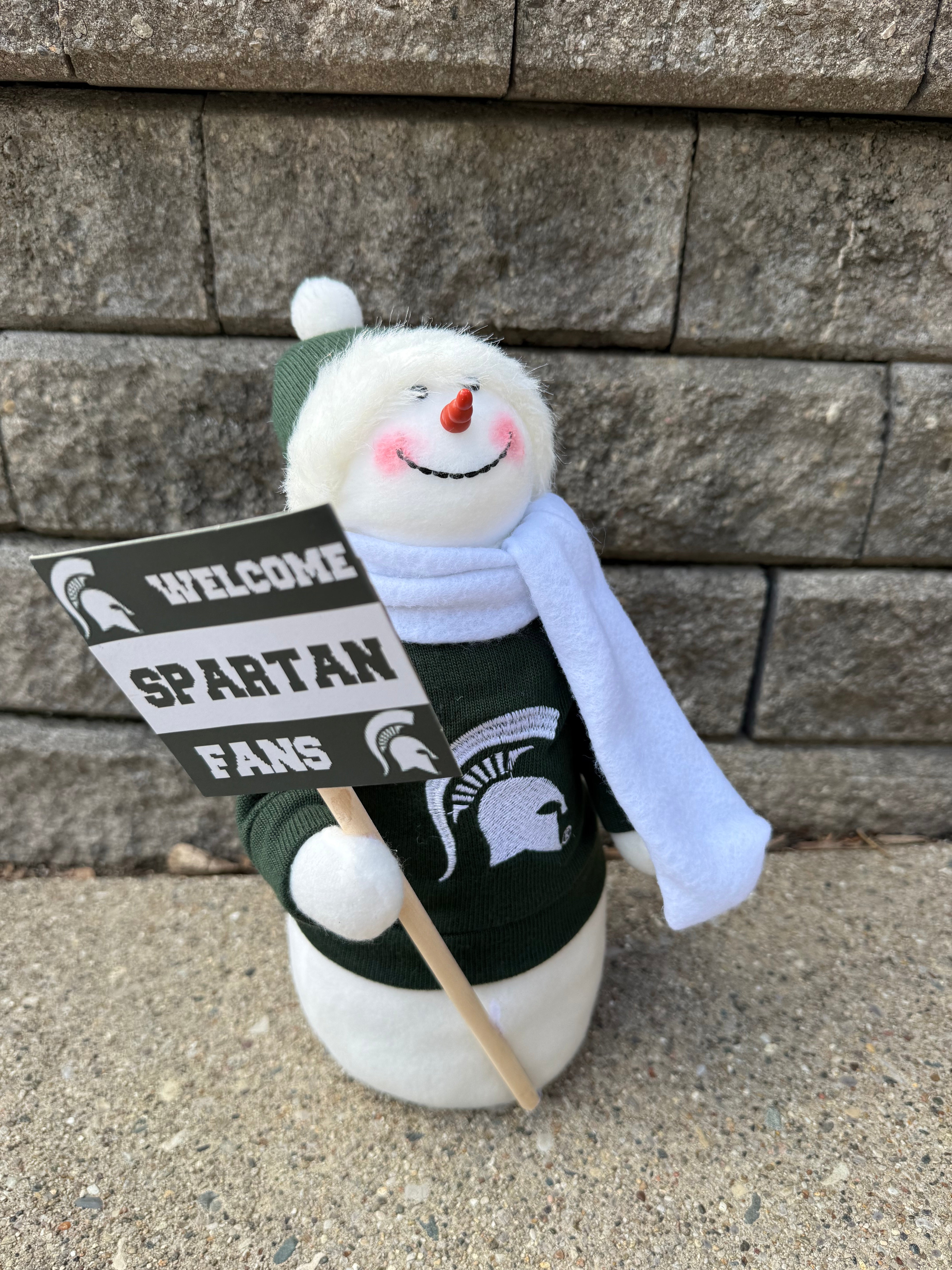 Snowman figurine wearing a winter hat, green sweater with a Spartan helmet logo on it, and a white scarf. The snowman is holding a sign that says "Welcome Spartan Fans".