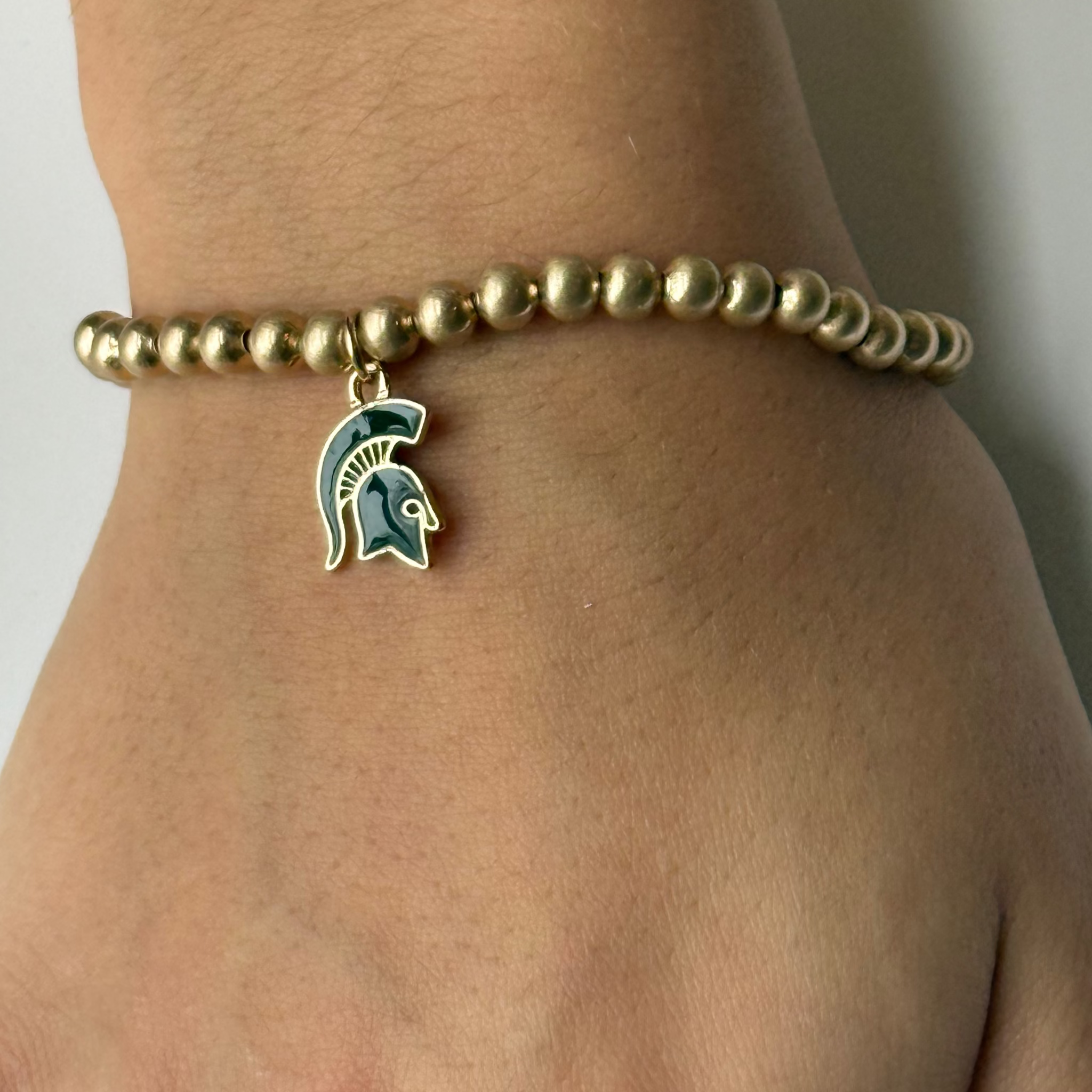 Close up of a gold beaded bracelet with a green and gold Spartan Helmet emblem. Shown on a wrist with a neutral background.