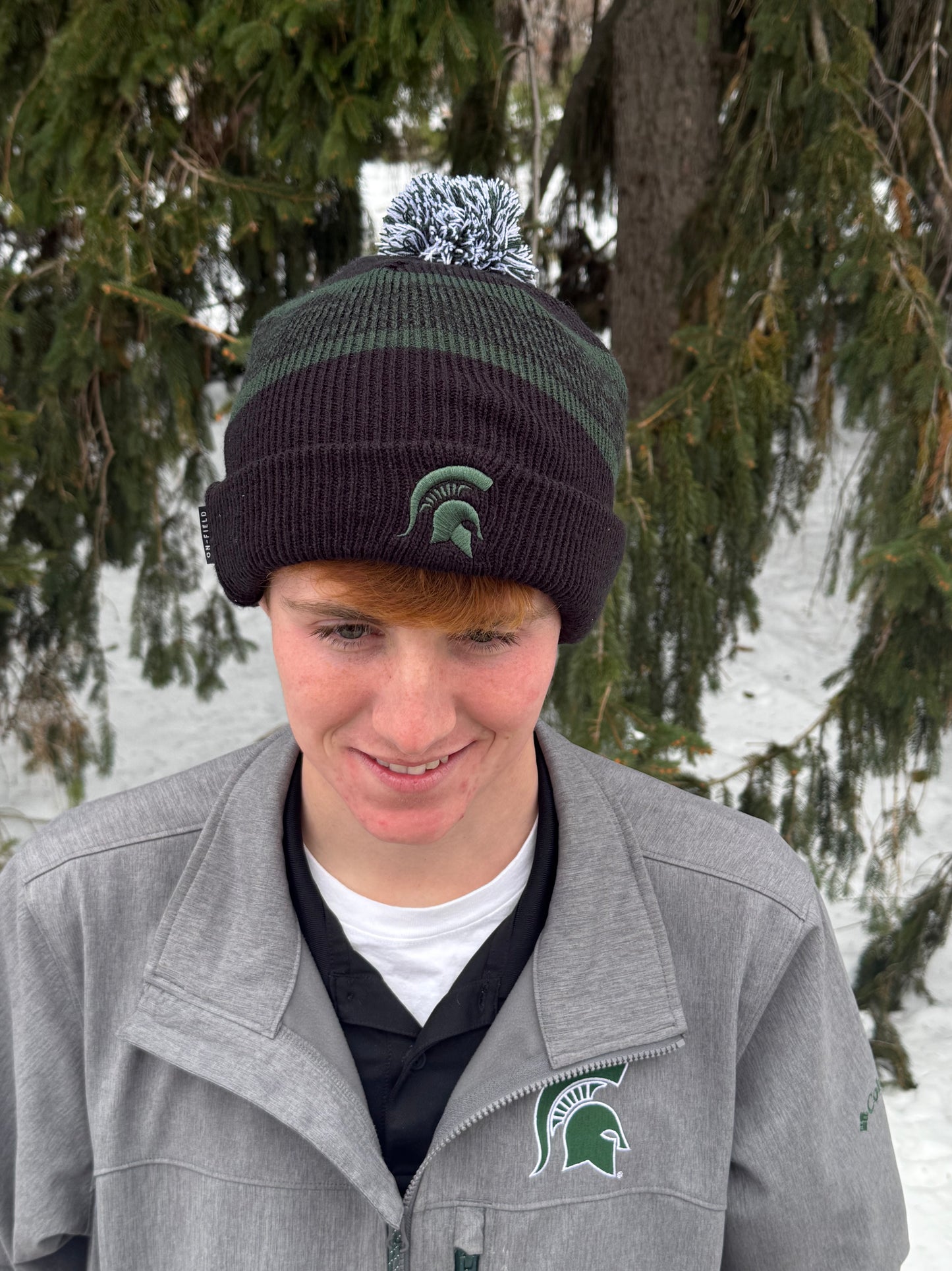 Man standing outdoors, wearing a black cuffed beanie with a white and green pom on the top, green stripe in the middle, and an embroidered green Spartan Helmet logo on the cuff.