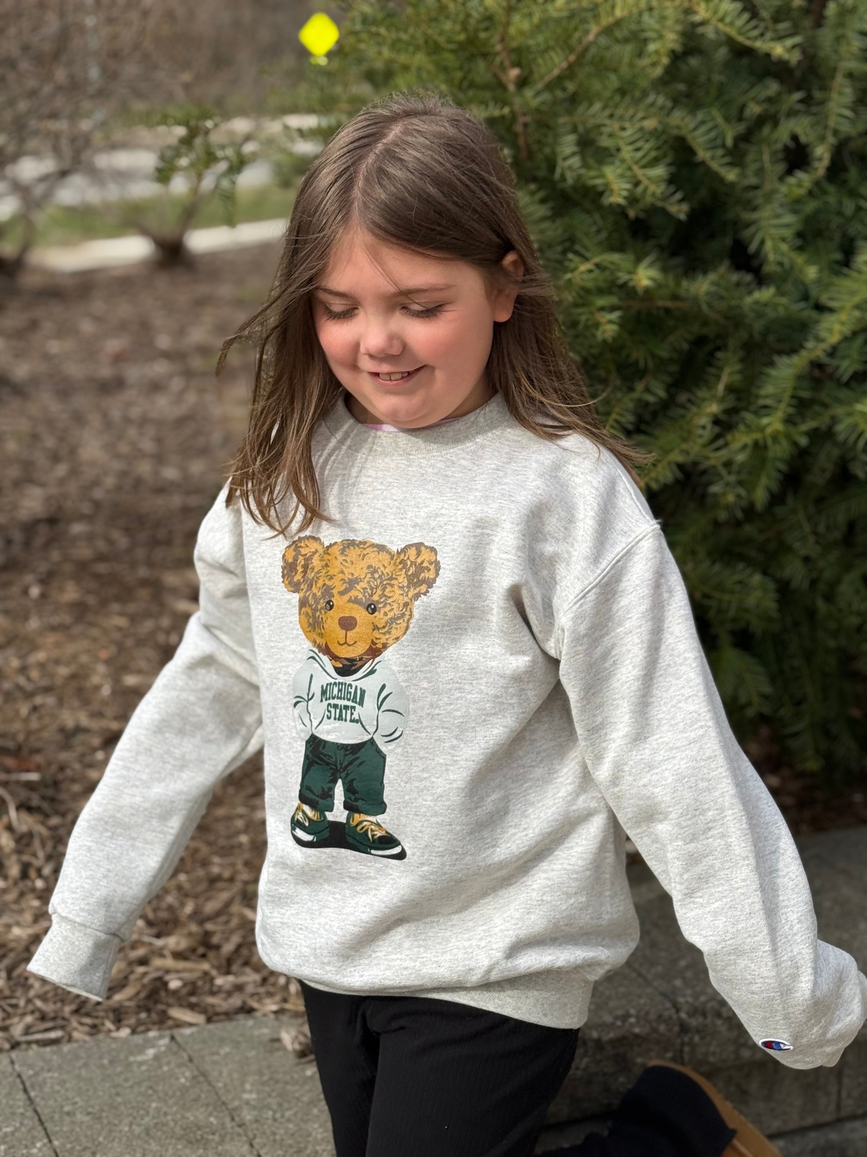 Child wearing a light beige crewneck sweatshirt with a teddy bear graphic that is wearing a sweatshirt with "Michigan State" written on it, green pants, and green and white shoes. The classic Champion logo is embroidered on the lower left cuff.