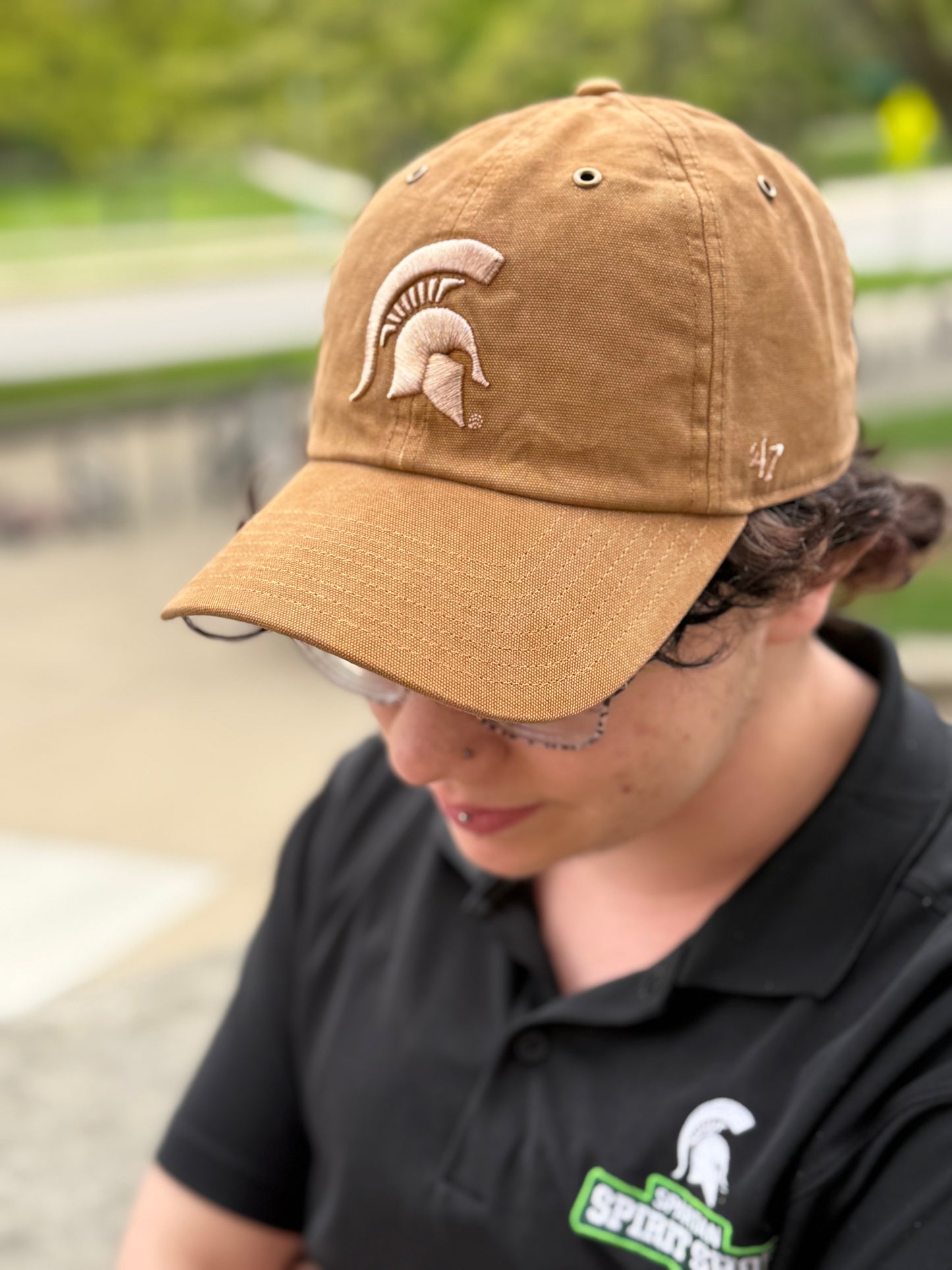 Person standing outdoors showing a beige hat with a light beige Spartan Helmet logo embroidered on the front.