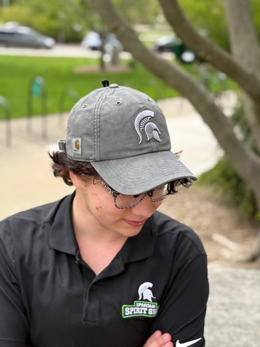 Person standing outdoors showing a gray hat with a light gray Spartan Helmet logo embroidered on the front and Carhartt logo patch on the right side.