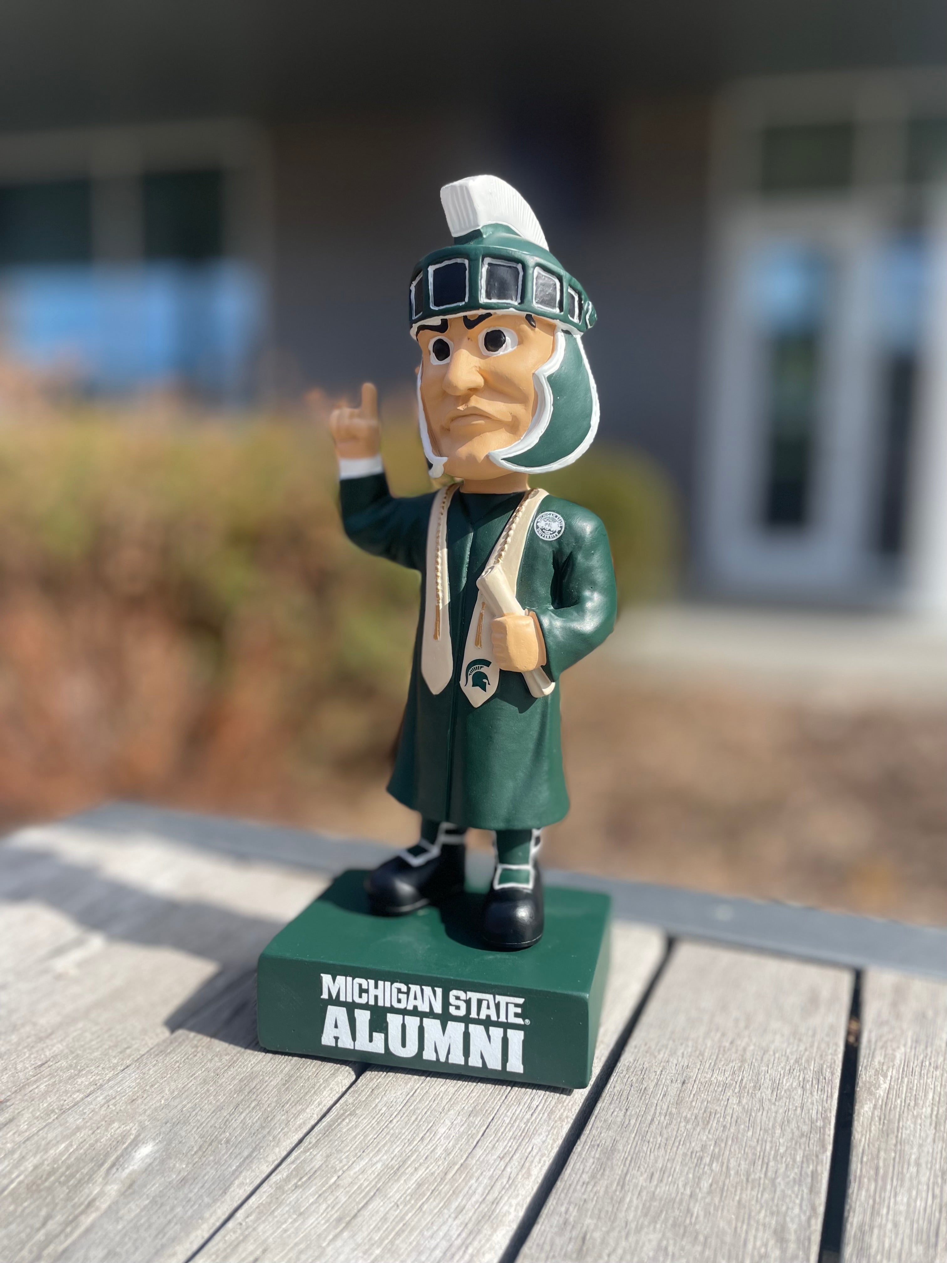 Graduation Sparty bobblehead wearing graduation attire and featuring bold text that reads "Michigan State Alumni" base.
