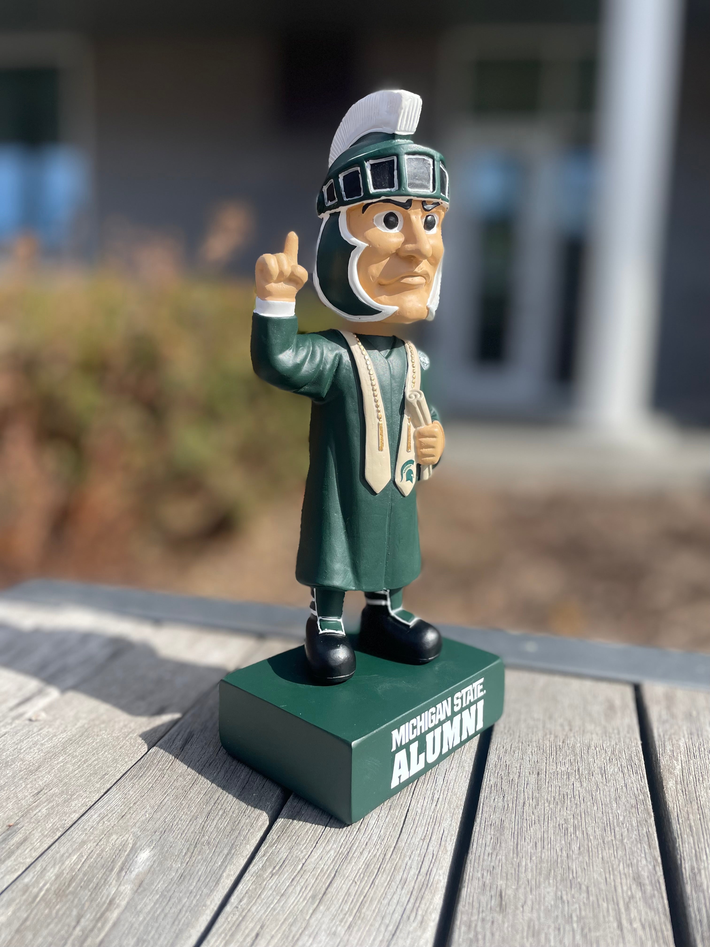 Side view of a graduation Sparty bobblehead wearing graduation attire and featuring bold text that reads "Michigan State Alumni" base.