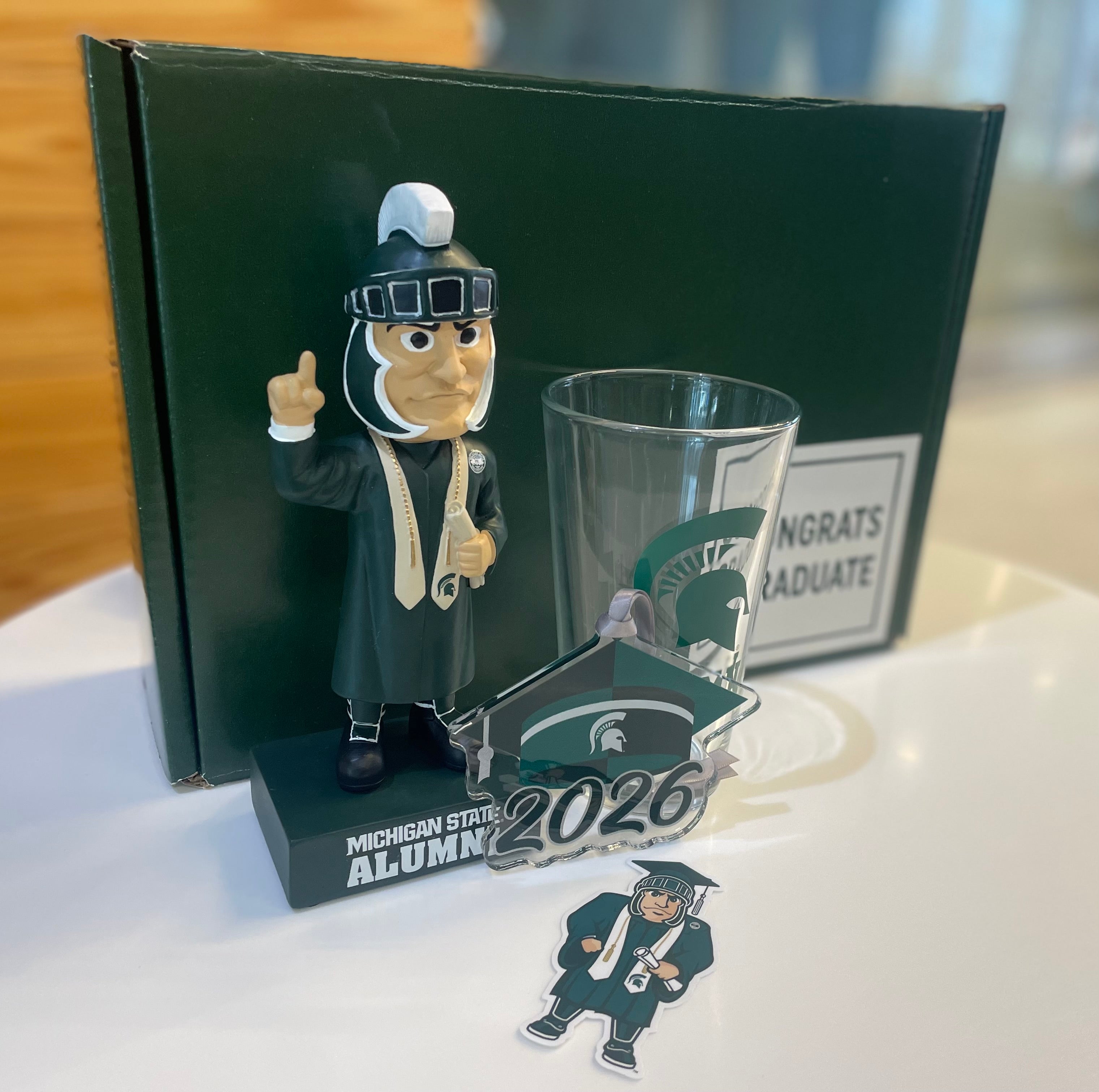 Graduation-themed set with a glass, bobblehead, Grad Sparty sticker, 2026 grad cap MSU ornament with a 'Congrats Graduate' green box.
