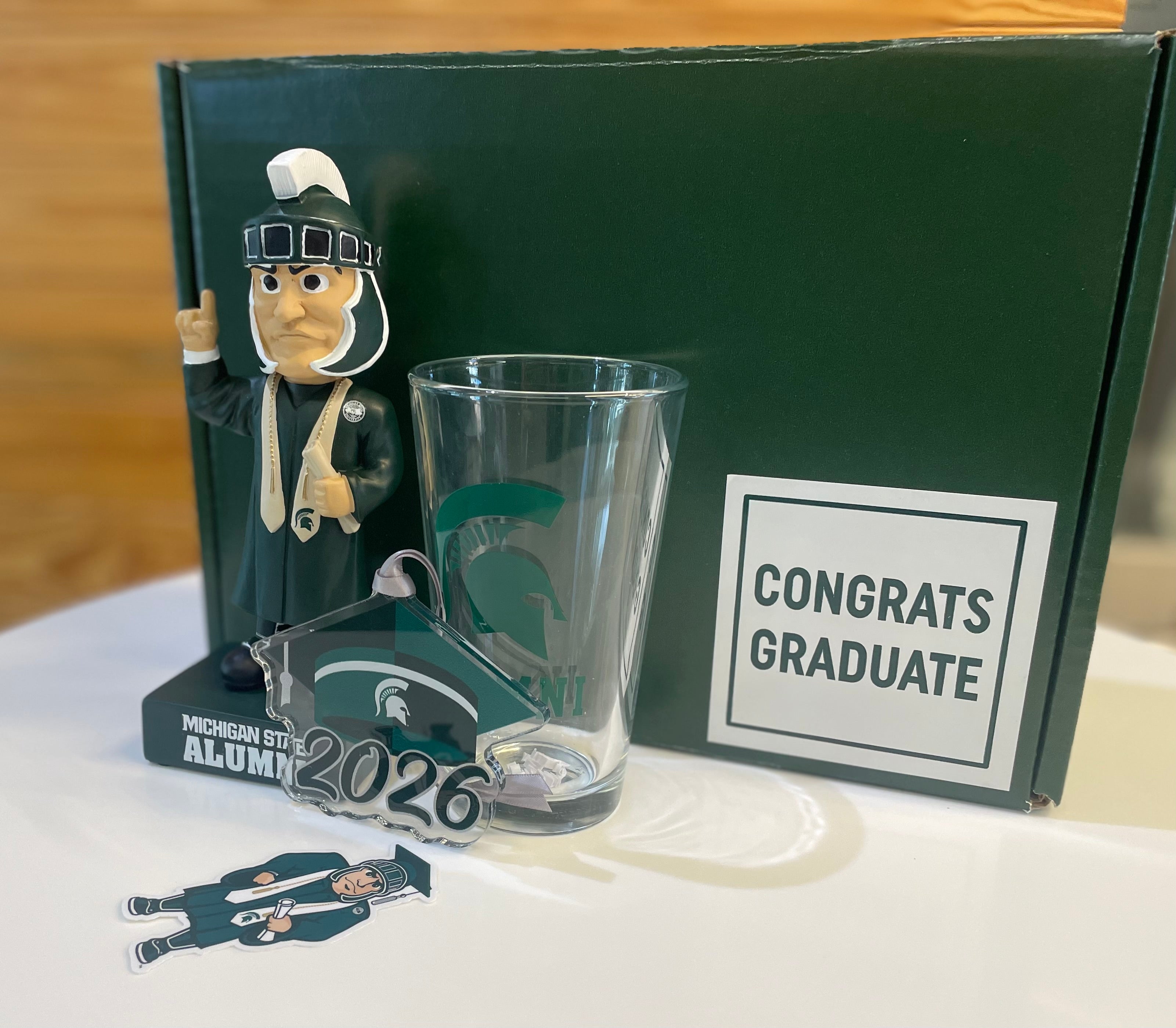 Graduation-themed set with a glass, bobblehead, Grad Sparty sticker, 2026 grad cap MSU ornament with a 'Congrats Graduate' green box.