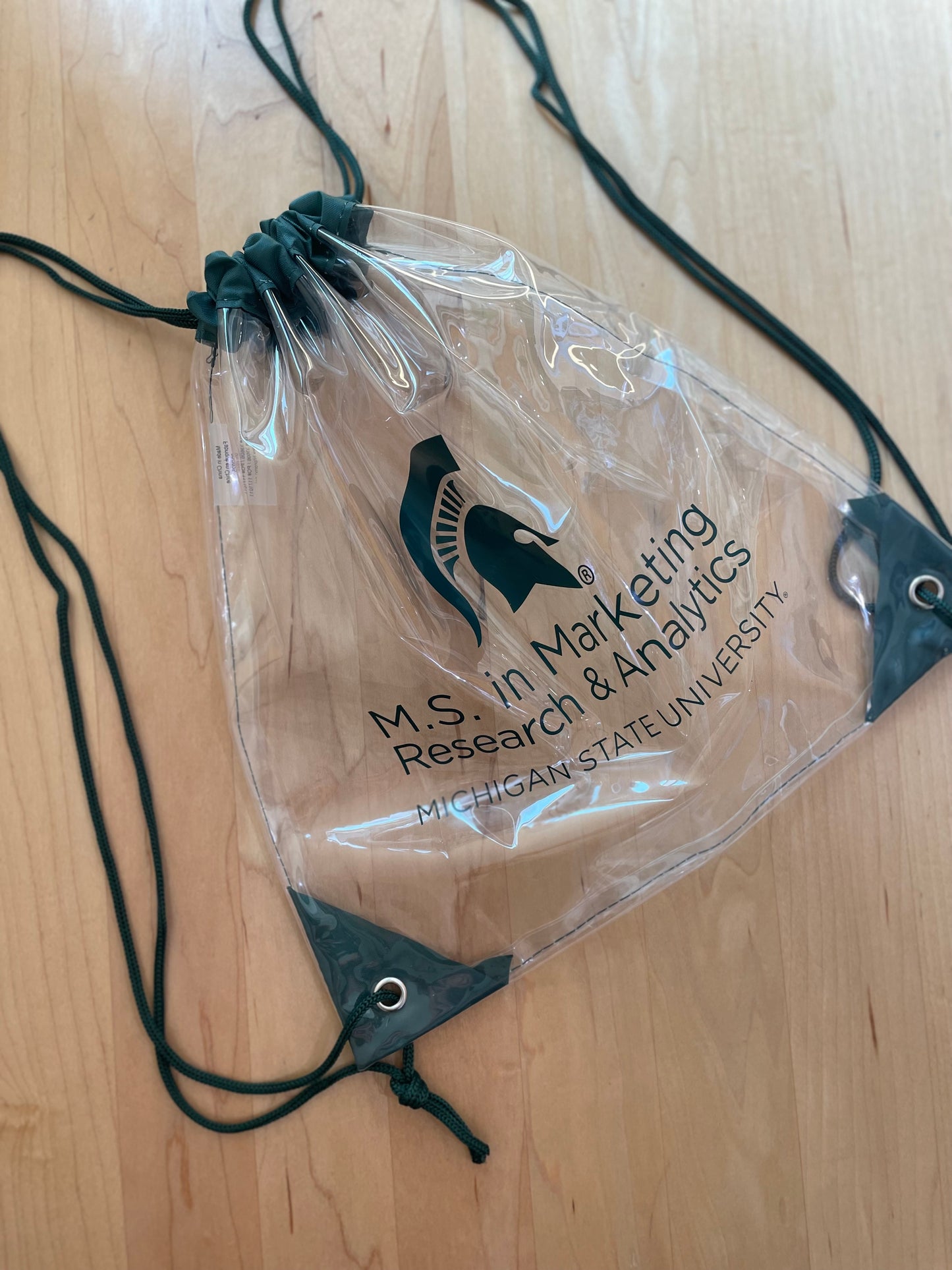 Cinched clear drawstring backpack with the M.S. in Marketing Research & Analytics Michigan State University logo printed in green. Green trim and string handles.