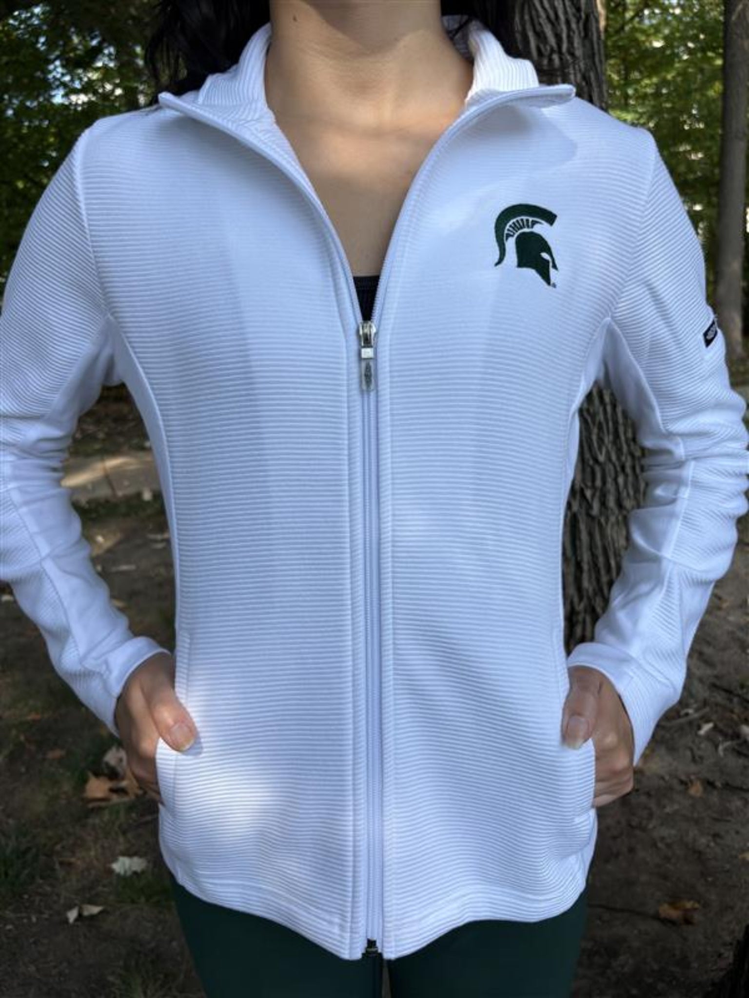 Person in a white long-sleeve hoodie jacket with a green embroidered Spartan helmet logo on the left chest.