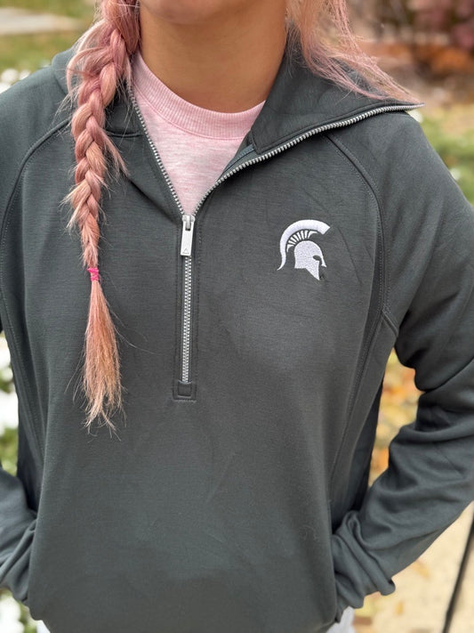 Close up of woman, standing outdoors, wearing a gray quarter-zip with a white Spartan Helmet logo on the left upper chest and an extended collar.